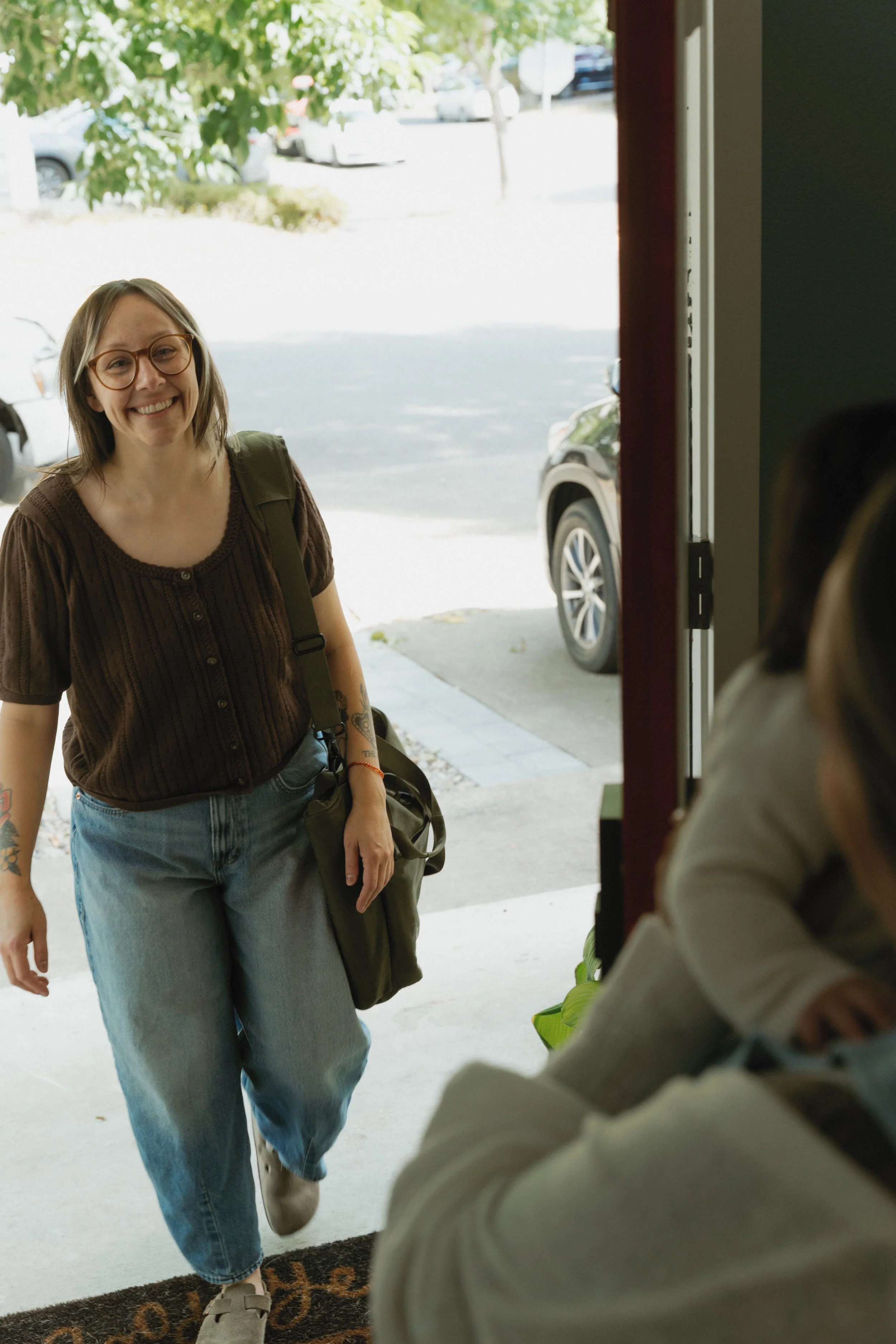 A woman with glasses and tattoos smiling as she enters a building, with parked cars and trees outside visible through the doorway.