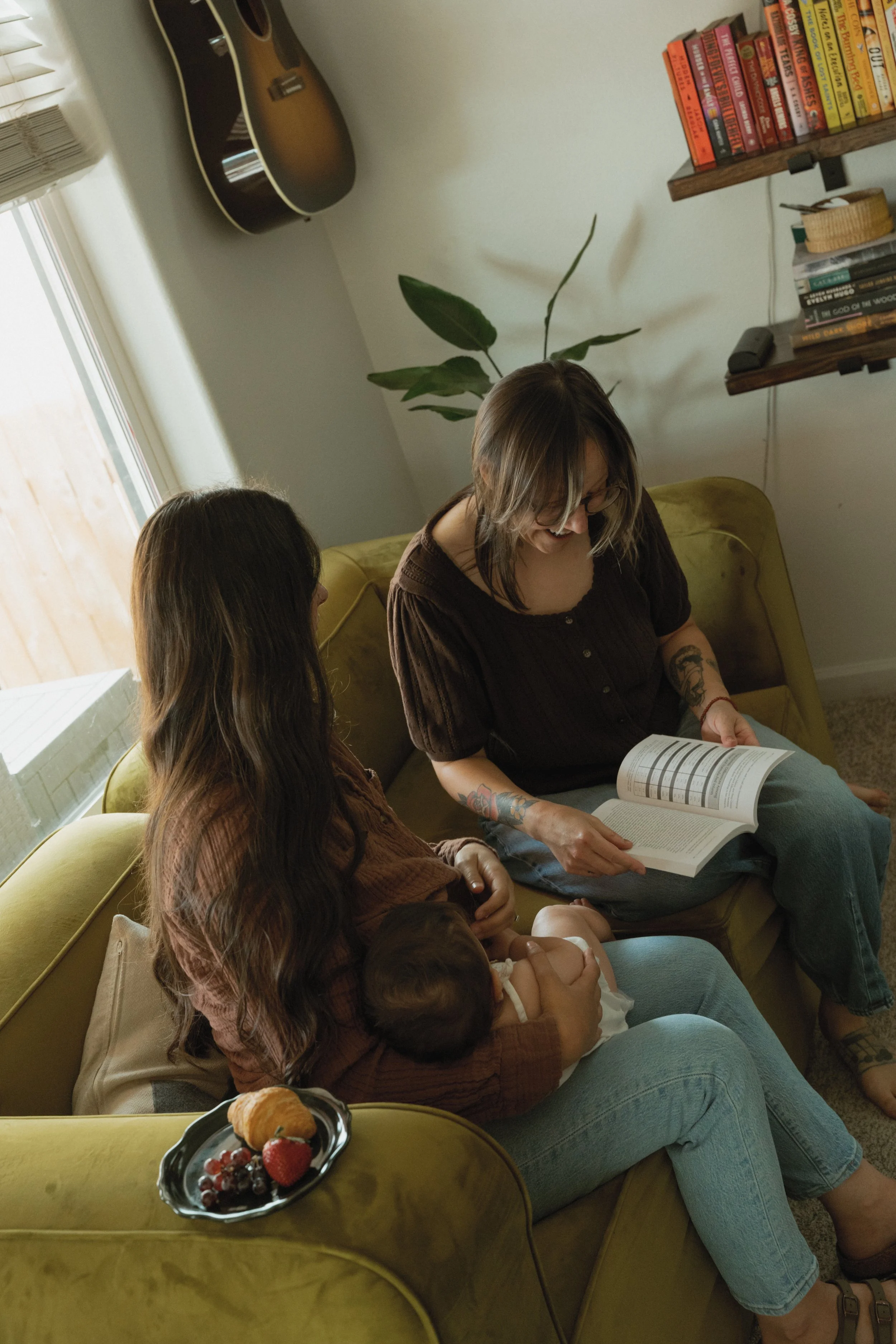 Two women are sitting on a yellow sofa in a cozy room, engaged in reading and conversation. One woman has glasses, tattoos, and is holding a book, while the other woman is listening attentively. On a table in front of them, there is a plate with strawberries, blueberries, and pastries. The room has bookshelves filled with books, a guitar hanging on the wall, a large plant, and an air conditioner near a window.