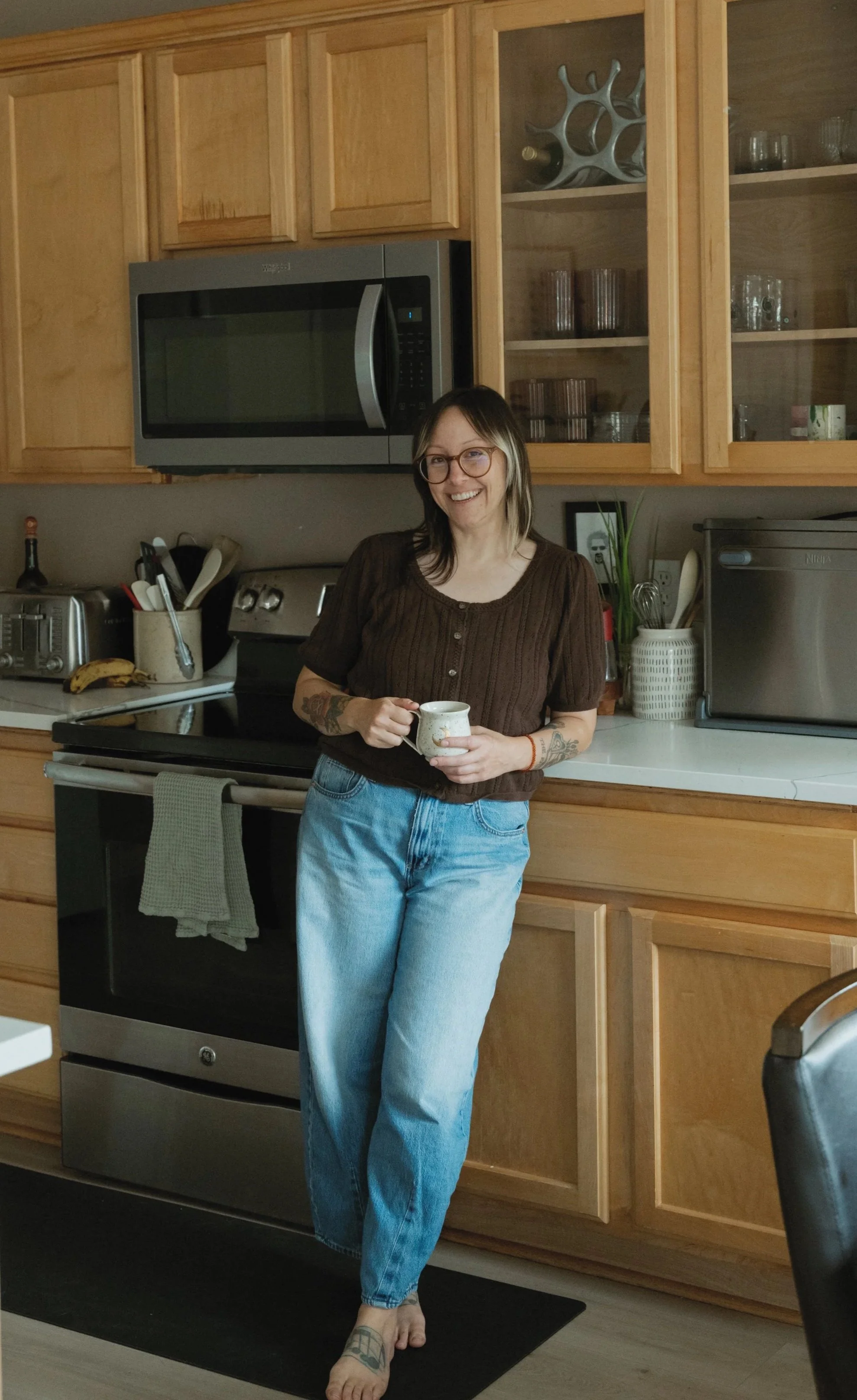 A woman with glasses, tattoos, and casual clothing standing in a kitchen, holding a mug and smiling.