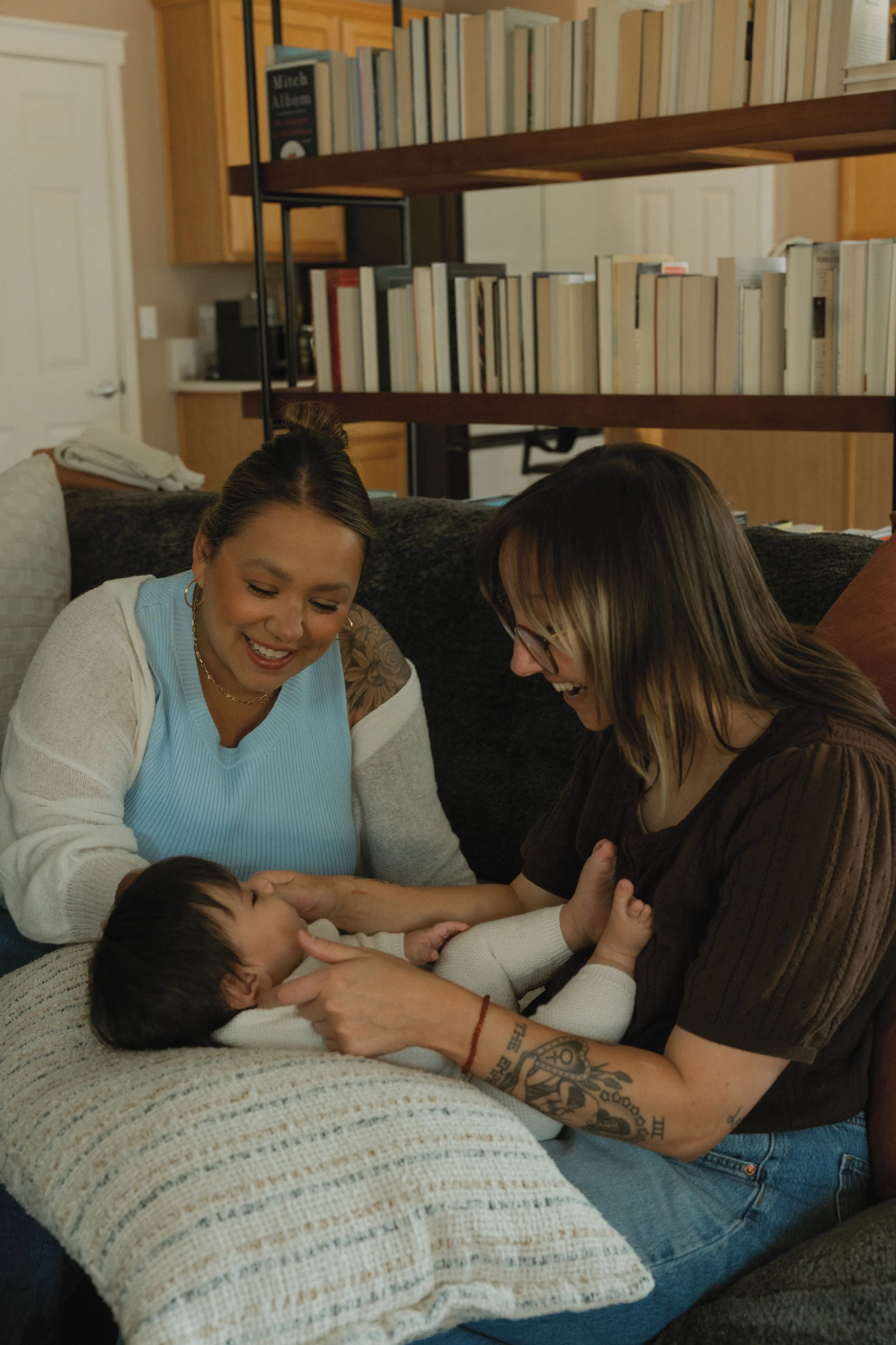 Two women and a young child are gathered on a couch, engaging in playful interaction and smiling. The setting is a cozy living room with bookshelves in the background.