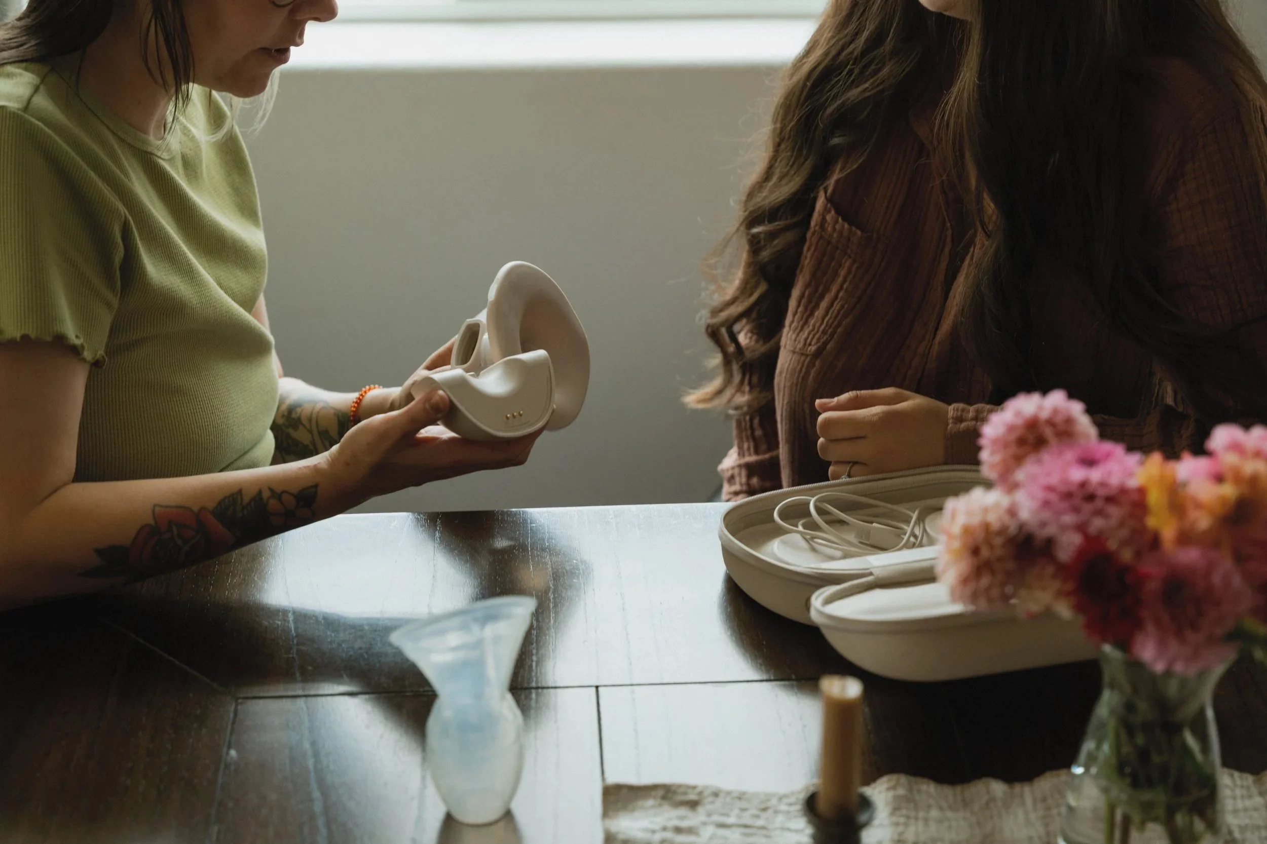 Two women sit at a table, one holding a white silicone model of an ear, with pink flowers in the foreground.