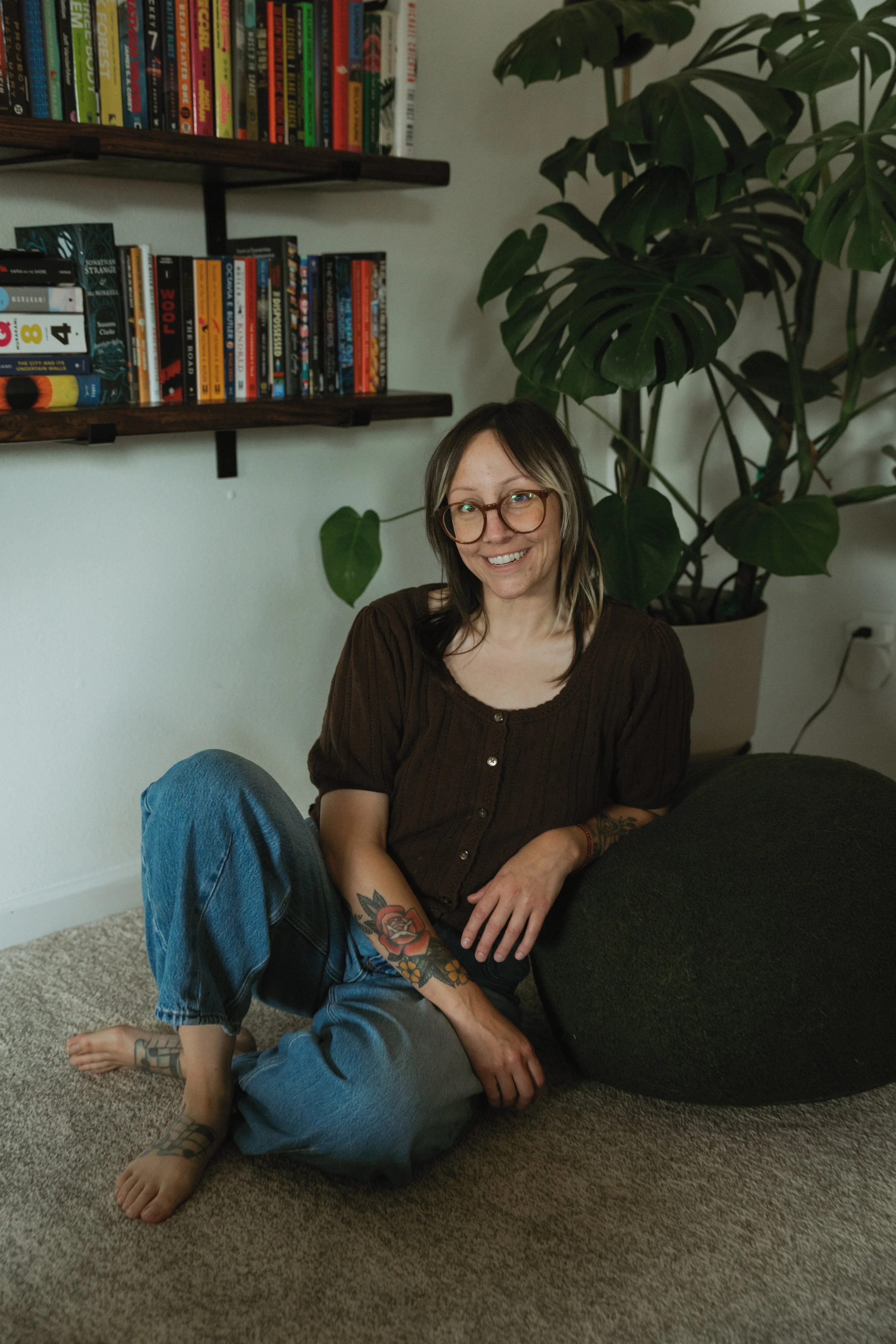 A woman with glasses and tattoos sitting on the floor next to a green couch, smiling at the camera. She is wearing a brown shirt and jeans, with a large plant in the background and books on wooden shelves behind her.