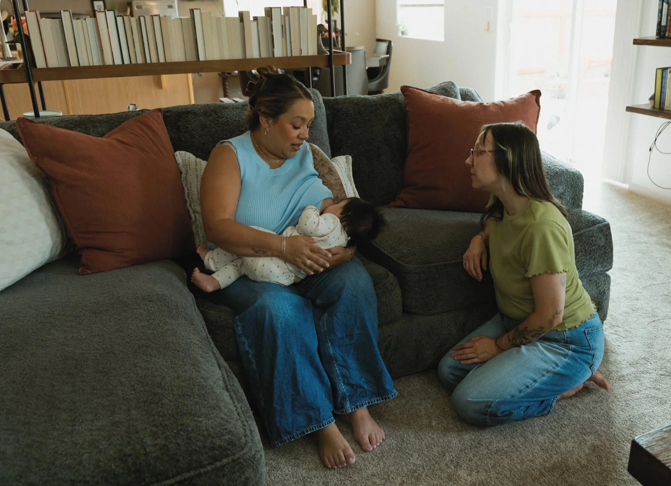 Two women are in a living room with a gray couch and red pillows. One woman with tattoos, wearing glasses and a yellow shirt, is kneeling on the carpet, looking at a baby. The other woman with dark hair, wearing a blue sleeveless top, is sitting on the couch holding the baby and looking at someone or something outside the frame.