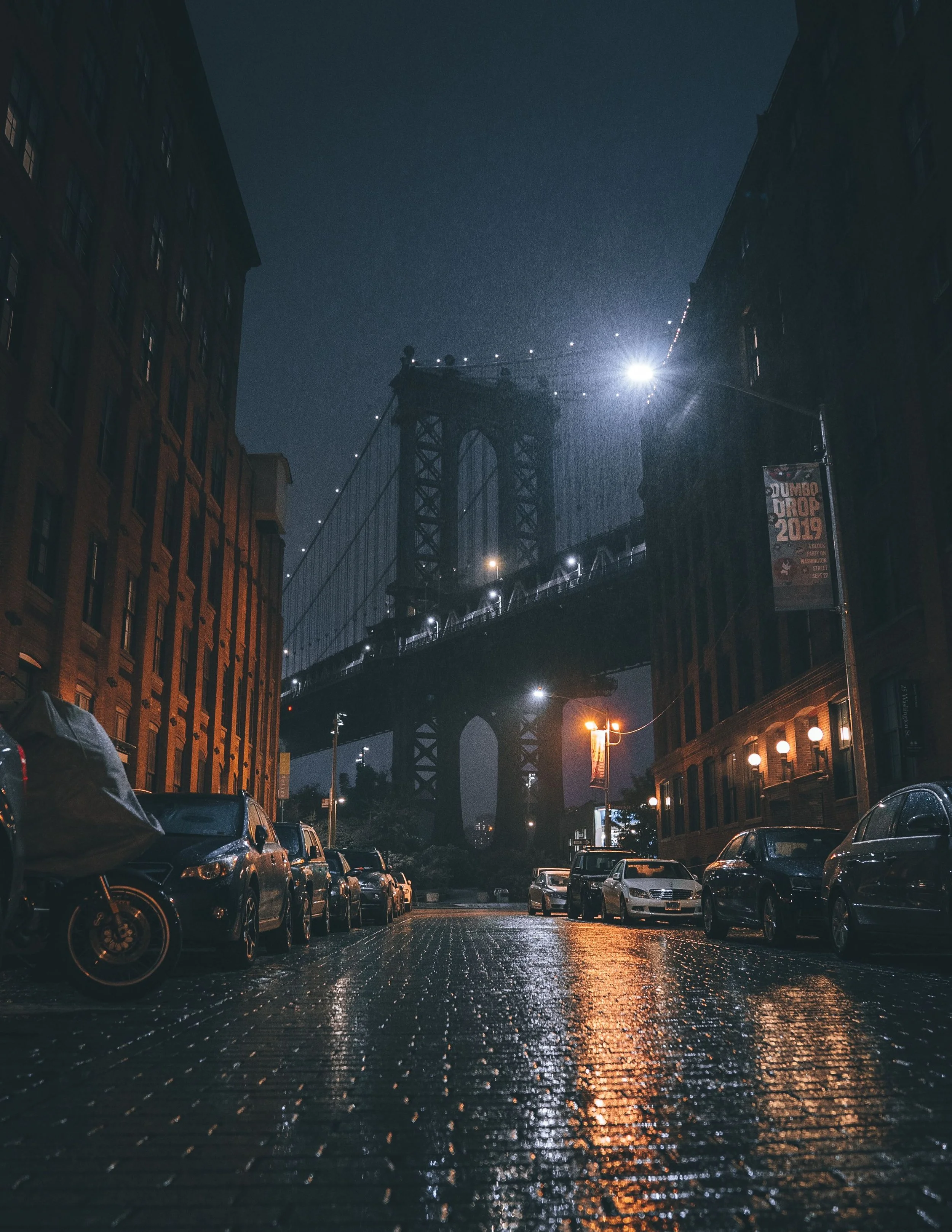 Nighttime street scene with parked cars, wet pavement reflecting streetlights, and a bridge in the background illuminated by spotlights.