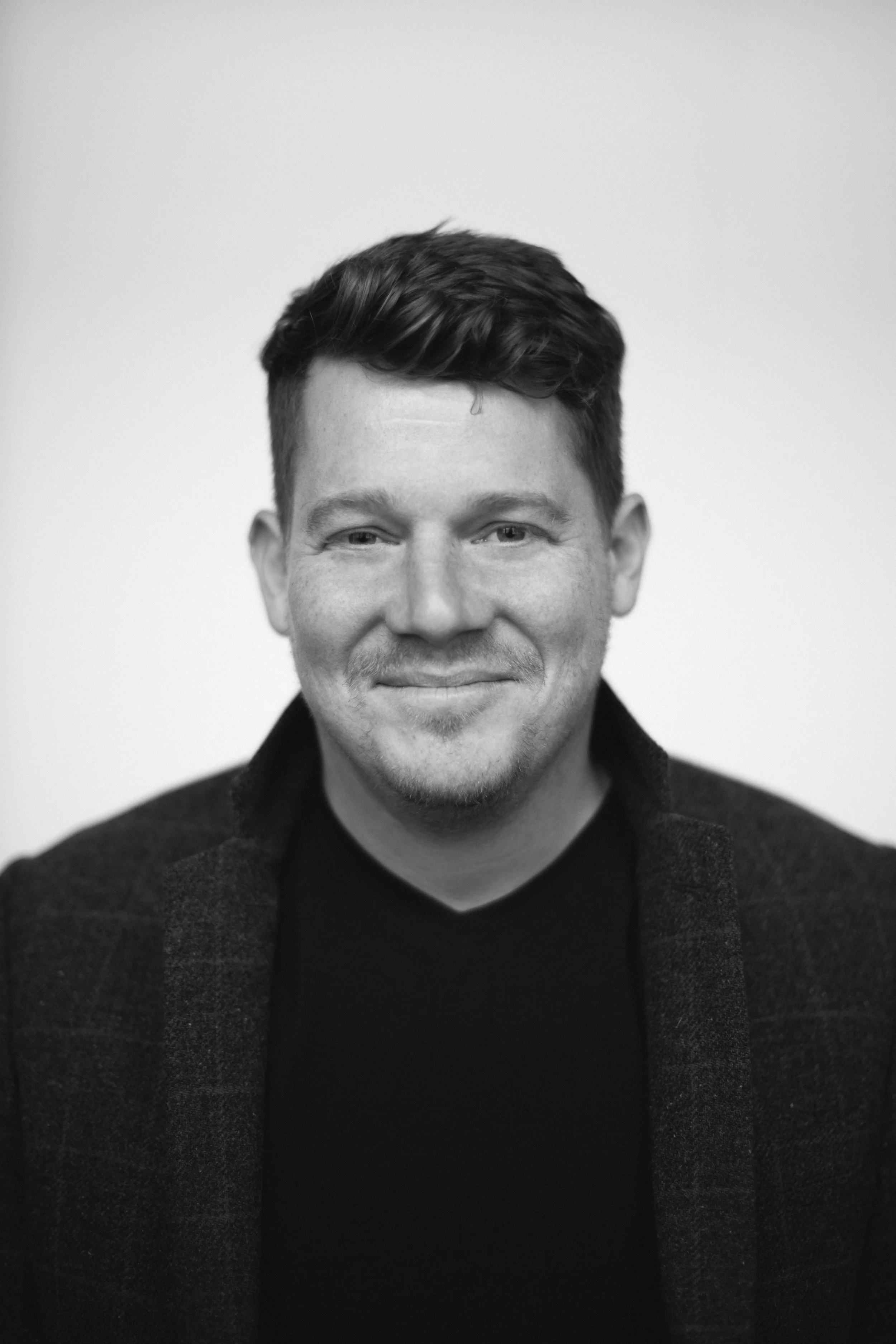 Black and white portrait of a smiling man with short, dark, wavy hair, wearing a dark turtleneck and a dark blazer, against a plain background.