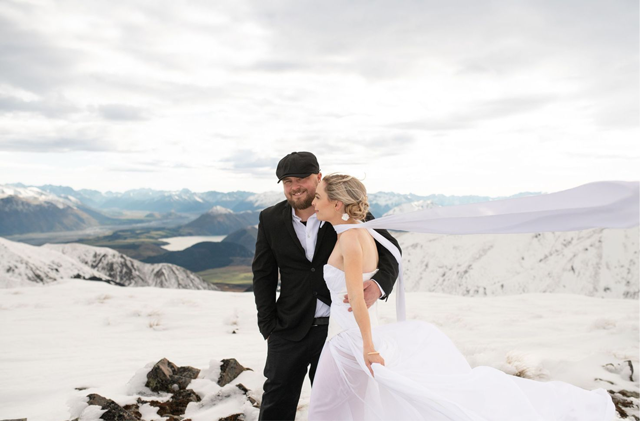 A smiling couple in wedding attire stands in a snowy mountain landscape. The bride is wearing a strapless white wedding dress with a long flowing train and a white scarf. The groom is in a black suit with a white shirt and a cap. They are embracing with mountains and a partly cloudy sky in the background.