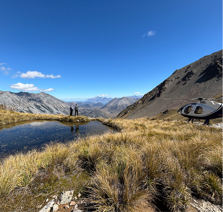 Two people standing near a small pond in a mountainous landscape under a clear blue sky, with a helicopter parked nearby.