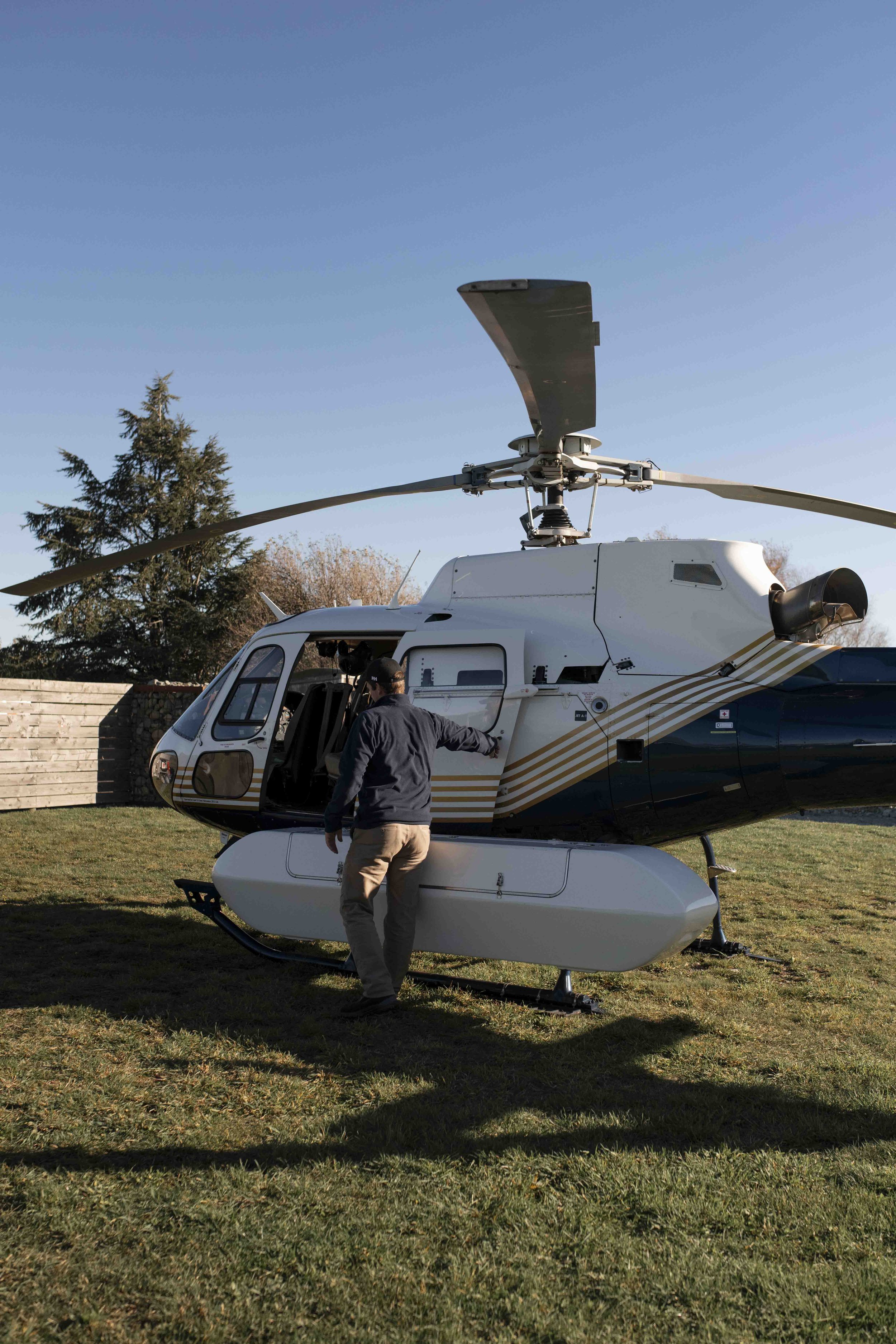 A helicopter with a person standing beside it on a grassy area, trees in the background, and a clear blue sky.