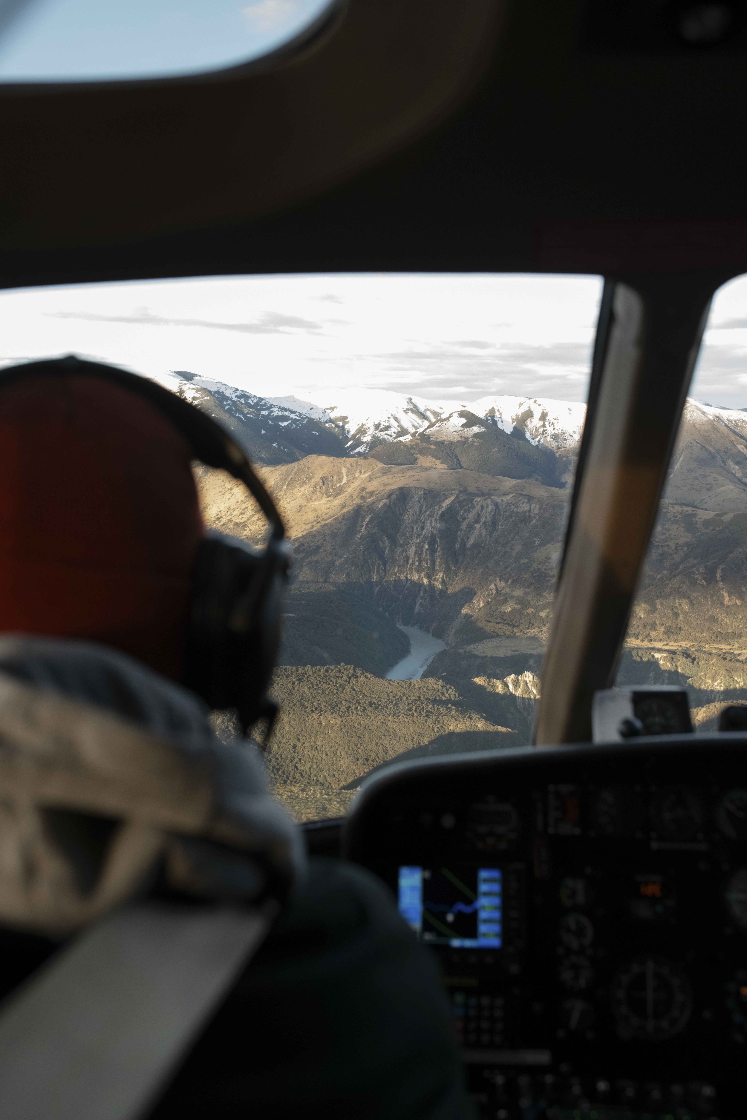 Inside a helicopter cockpit with a view of snow-capped mountains and a river below.