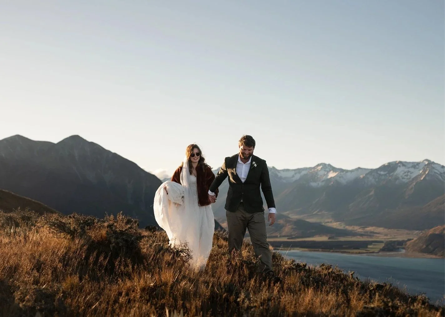 A couple in wedding attire walking hand in hand through a grassy landscape with mountains and a river in the background.