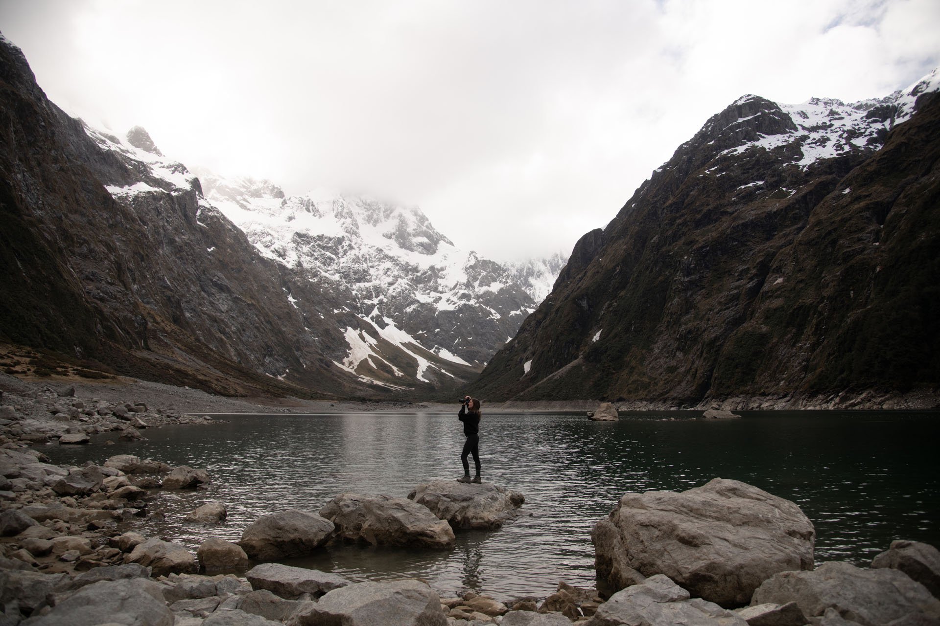 A person standing on rocks near a lake in a mountainous landscape with snow-covered peaks and cloudy skies.