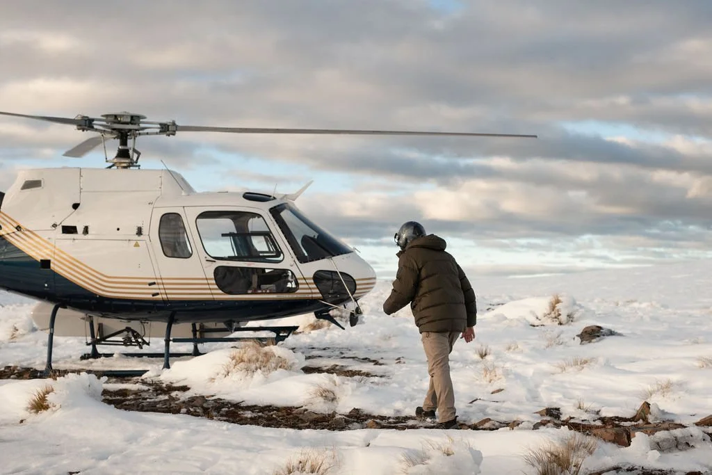 A person wearing a helmet and winter clothing walking near a helicopter on a snow-covered landscape, with a cloudy sky overhead.