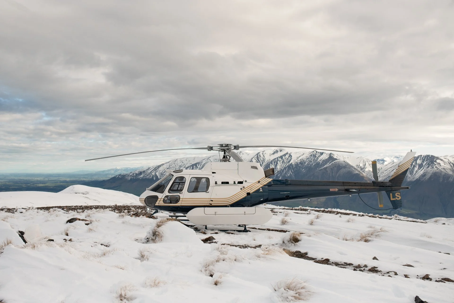 A helicopter on snowy ground near mountains with snow-capped peaks and cloudy sky.