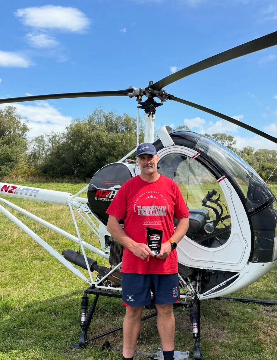 A man standing in front of a small white helicopter on a grassy field, holding a plaque or award, with trees and a blue sky in the background.