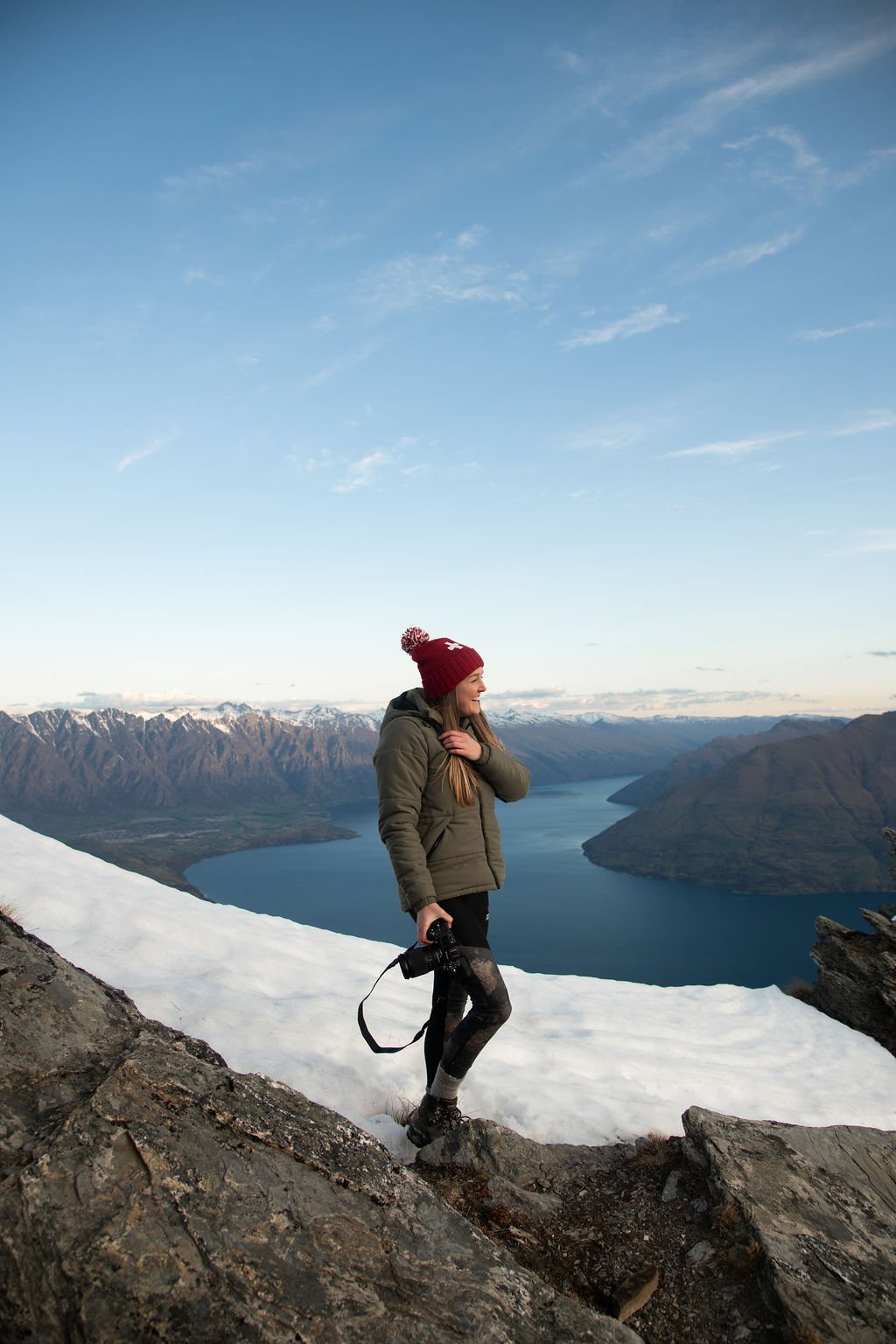 Woman hiking on snowy mountain trail with lake and mountain range in the background, wearing a red beanie, green jacket, and holding a camera.