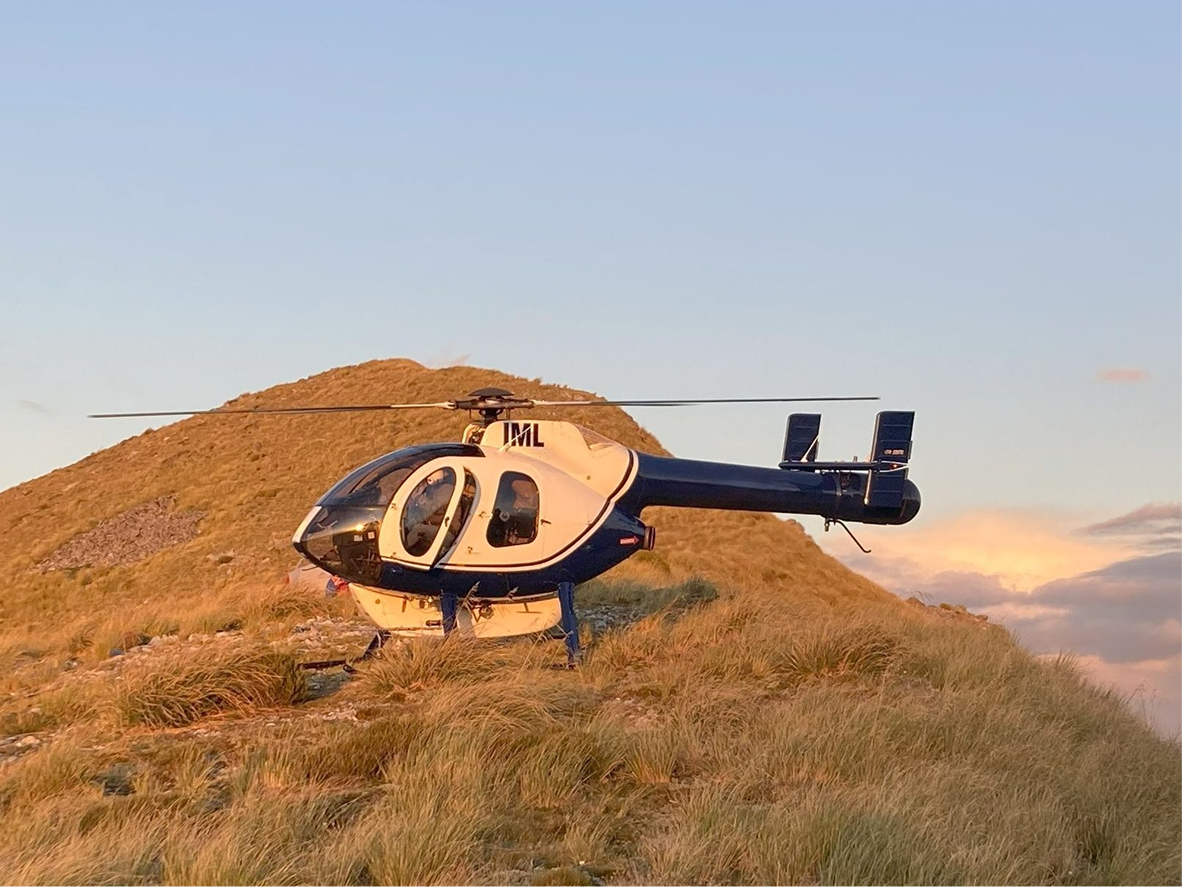 A helicopter on a grassy hillside with a mountain in the background during a sunset or sunrise.