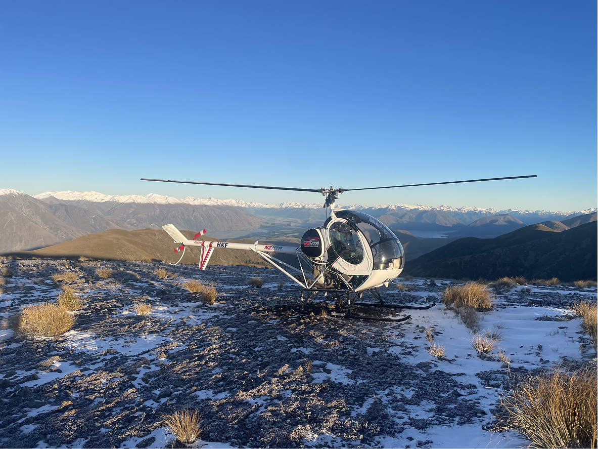 A helicopter on a rocky, snow-dusted mountain terrain with a scenic mountain range and snow-capped peaks in the background.