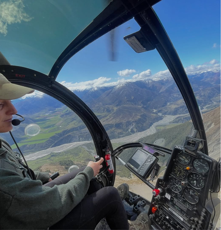 Pilot in helicopter cockpit flying over mountainous landscape with river and green fields.
