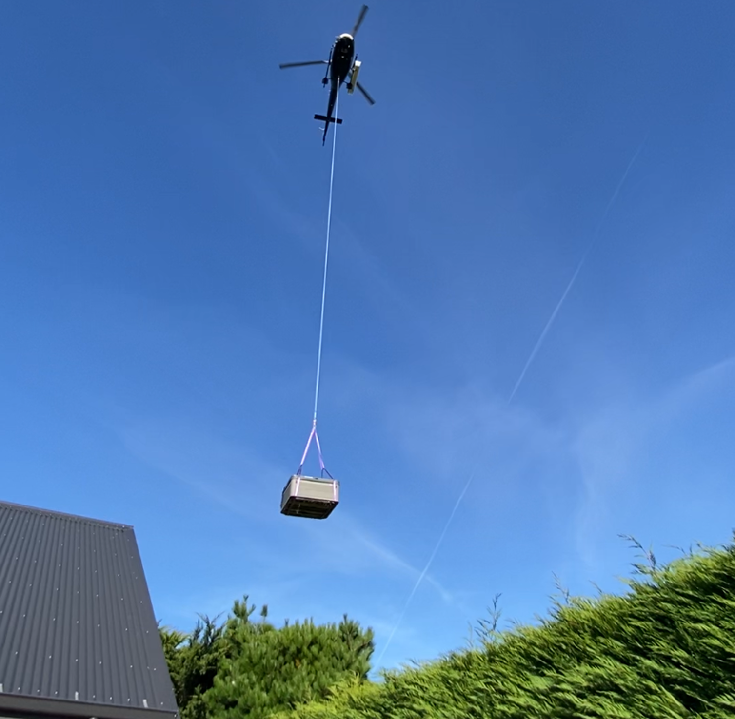 A helicopter is flying in the clear blue sky, towing a large container with a long cable. There are trees and a building with a dark sloped roof in the foreground.