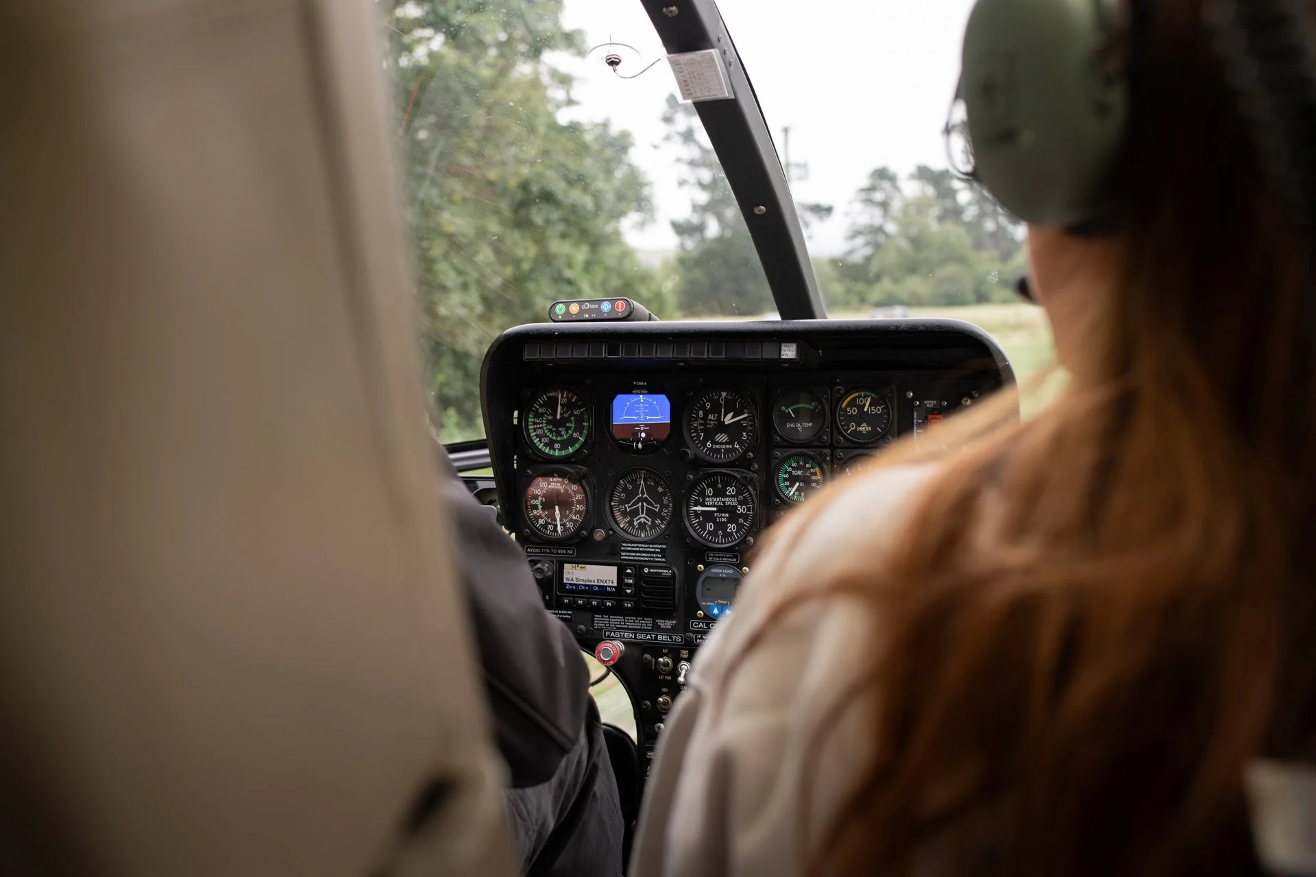 View of an airplane cockpit from the passenger seat, with two crew members visible, one wearing a headset. The control panel has various gauges and instruments, and outside the windshield, a cloudy sky and trees are visible.