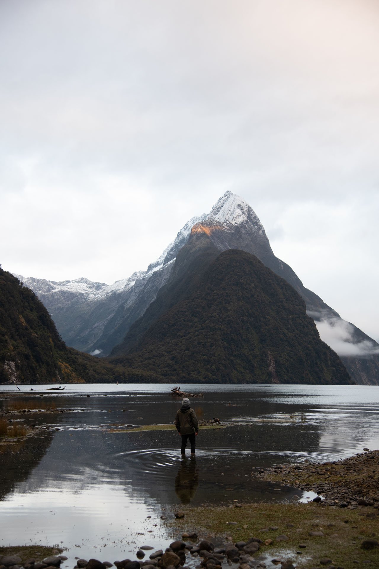 A person standing in shallow water on a rocky shore, with mountains with snow on top in the background and a lake in the foreground.