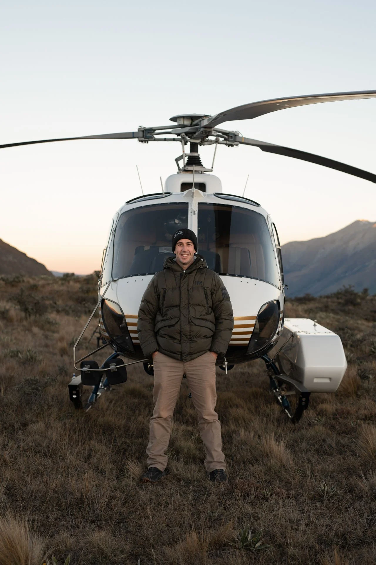 A man dressed in a brown jacket, beige pants, and a black beanie standing in front of a helicopter in a desert landscape with mountains in the background during sunset.
