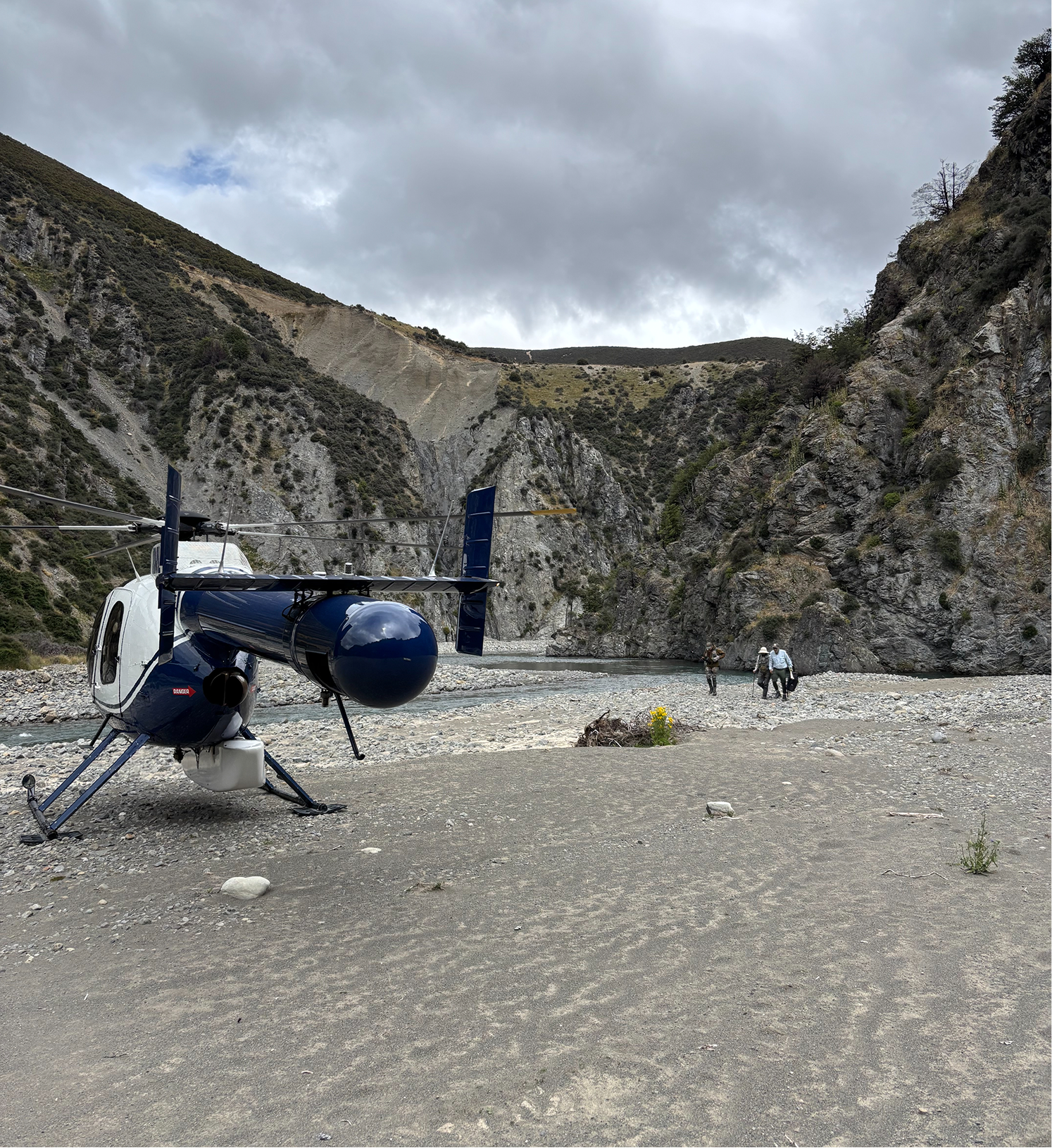A helicopter parked on rocky terrain next to a river, with a canyon and cloudy sky in the background. Two people are walking near the river.