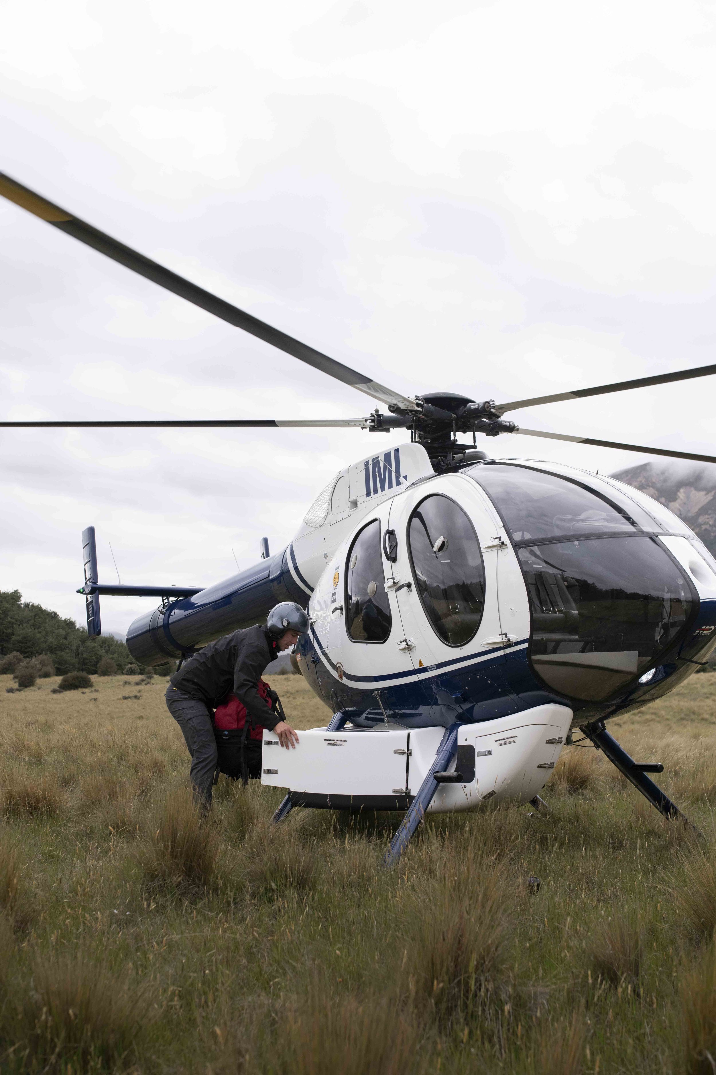 A helicopter is on the ground in a grassy field with mountains in the background. A person wearing a helmet and jacket is handling equipment near the helicopter.