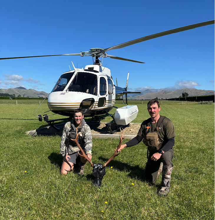 Two men in outdoor gear holding onto a set of antlers, kneeling on a grassy field in front of a helicopter with mountains in the background.