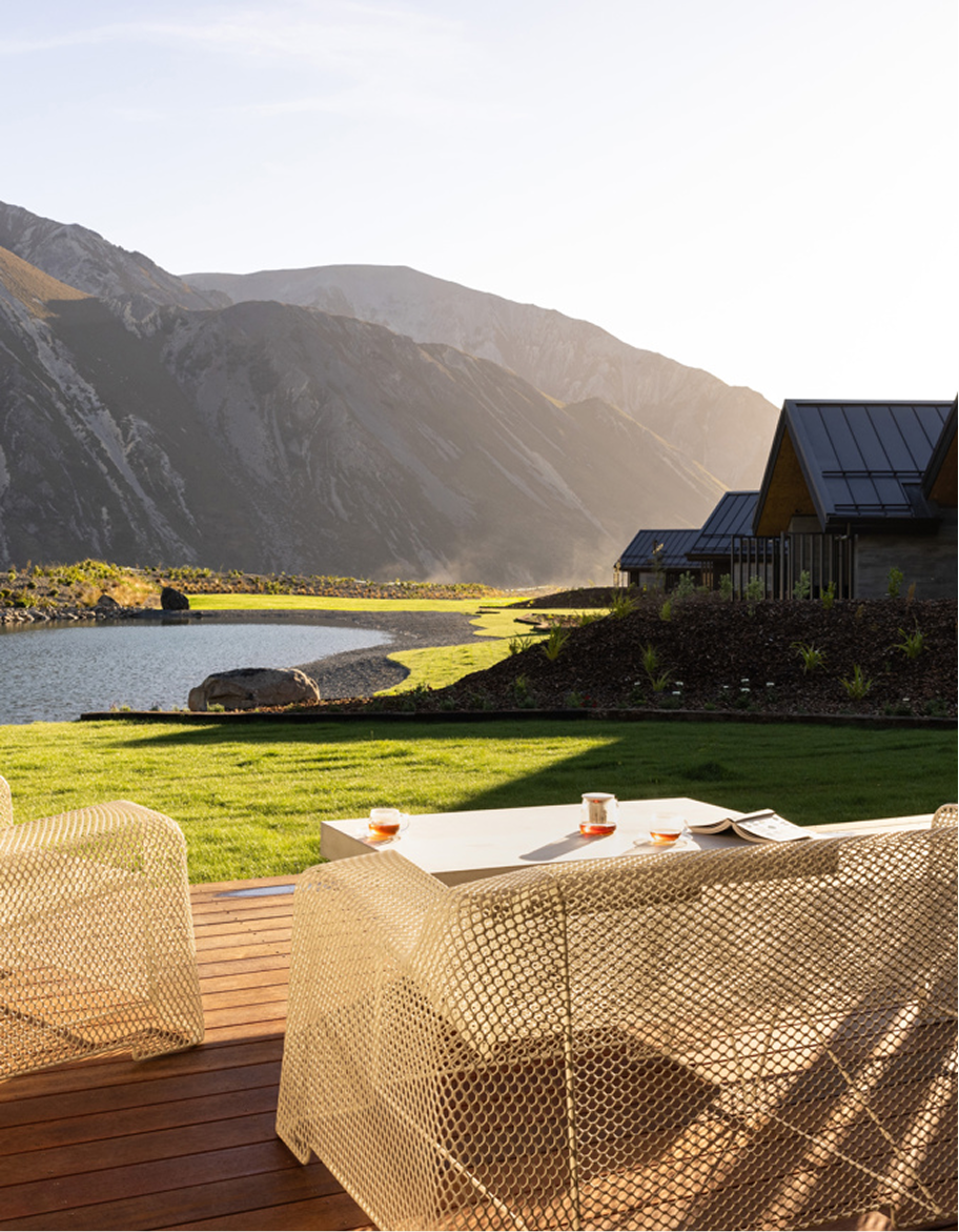 An outdoor seating area with a table and chairs on a wooden deck, overlooking a grassy yard, a lake, and mountains under a clear sky with sunlight.