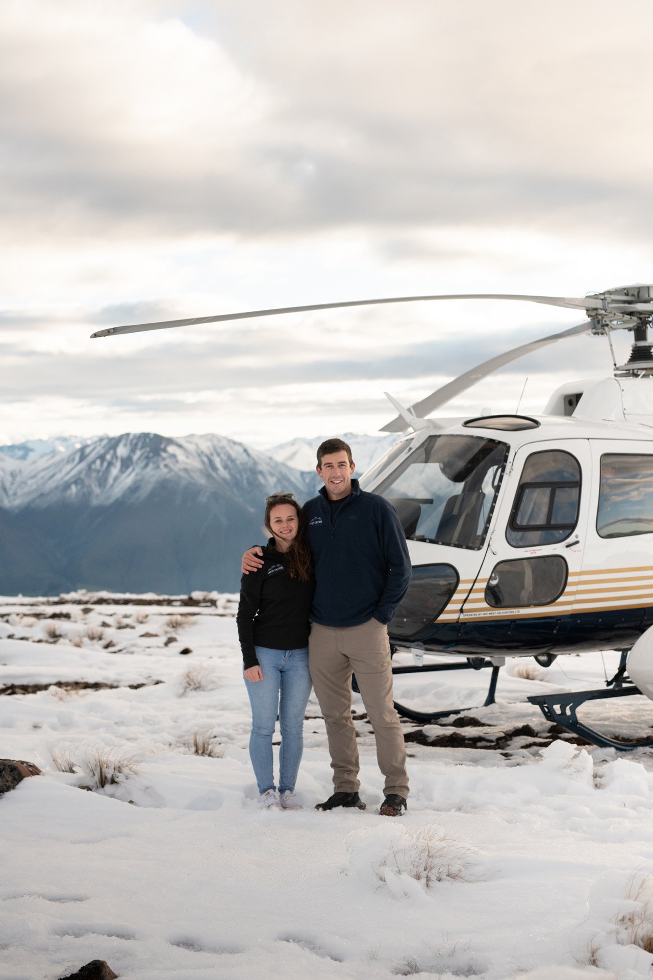 A man and woman standing together in snowy terrain near a helicopter, with mountains and cloudy skies in the background.