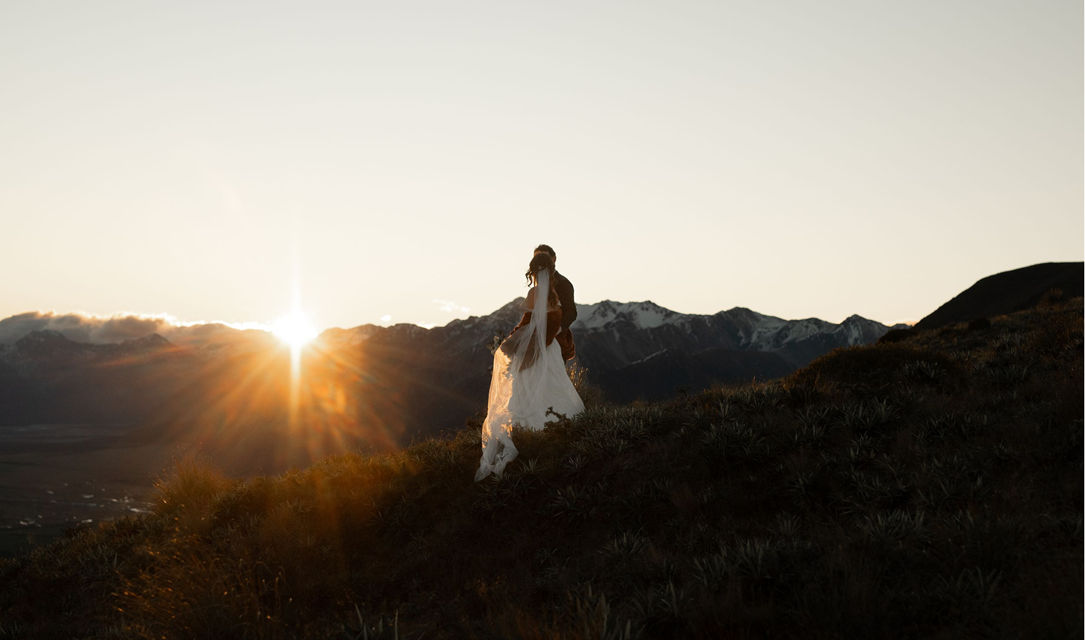 A couple standing on a grassy hillside at sunset, with mountains in the background.