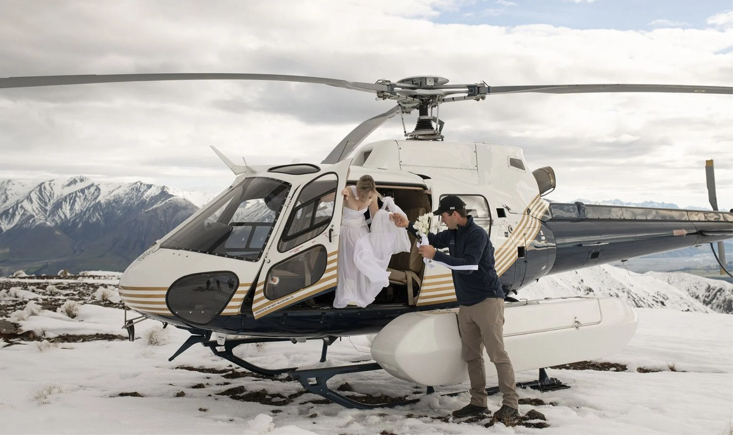 A couple getting married next to a helicopter in a snowy mountainous landscape. The bride is stepping out of the helicopter, and the groom is handing her a bouquet of white flowers.