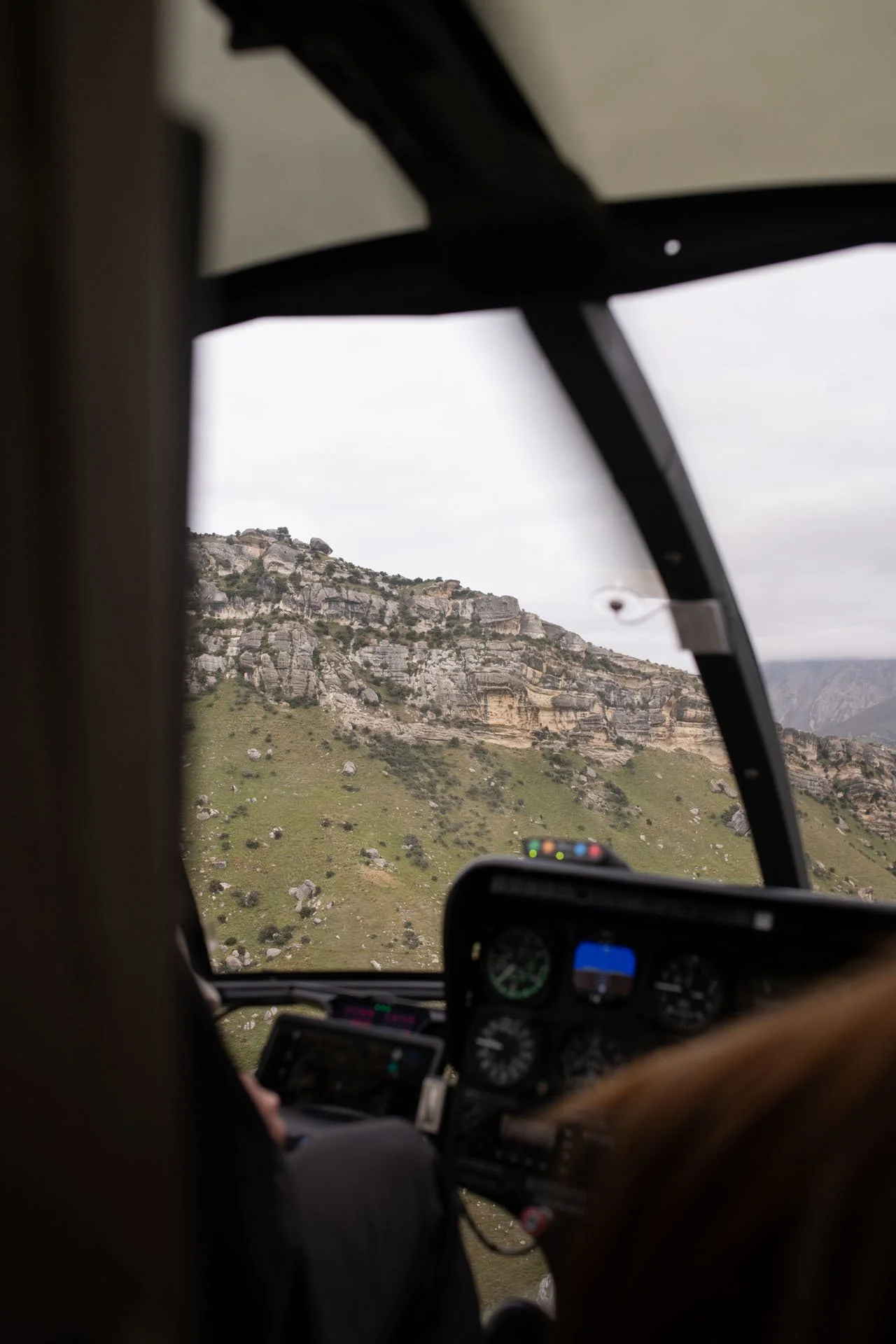 View from inside a helicopter cockpit showing the pilot's controls and dashboard, with a mountainous landscape visible through the window.
