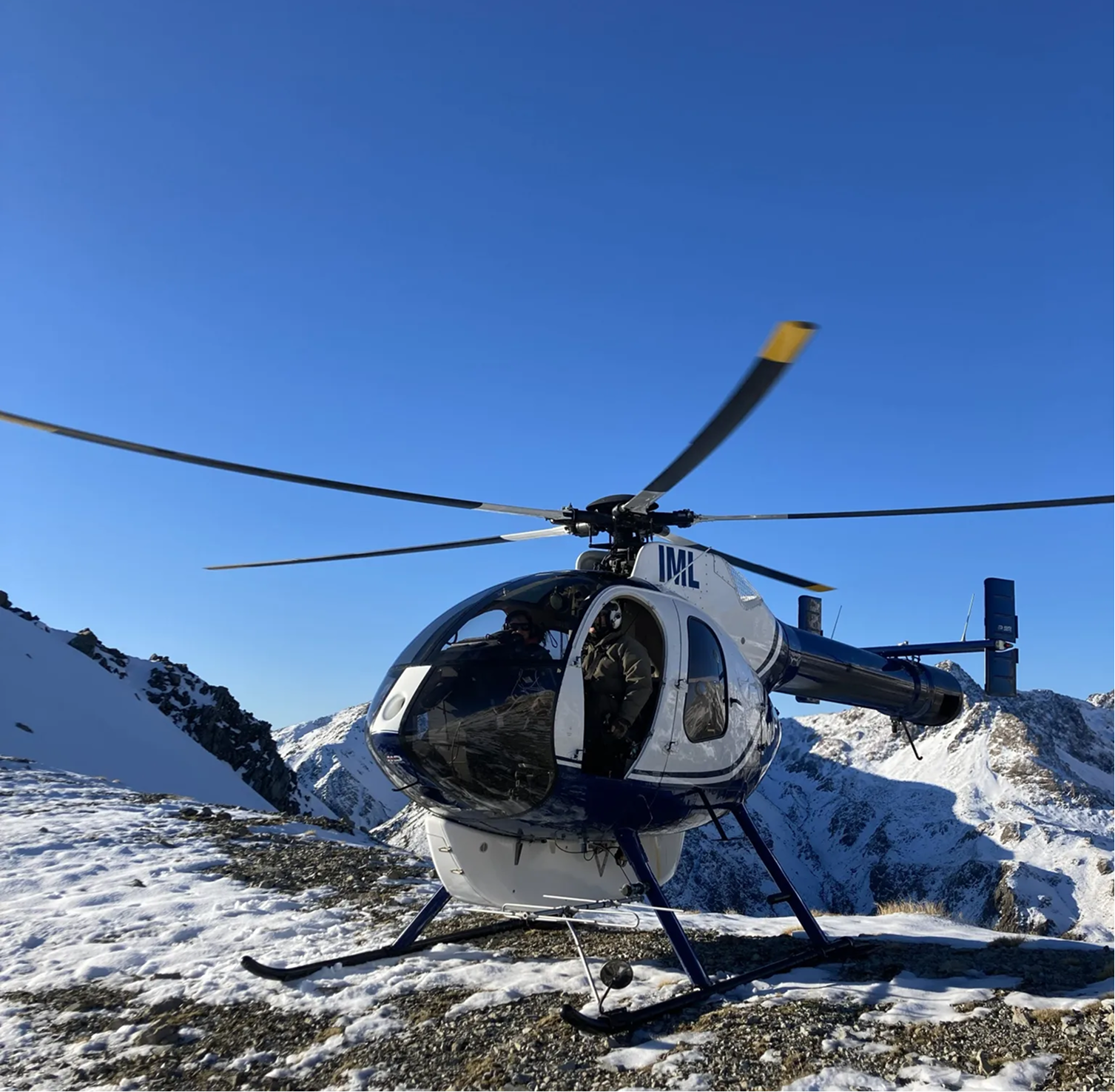 A helicopter, white and blue, landing on a snowy mountainous terrain with snow-covered peaks in the background under a blue sky.
