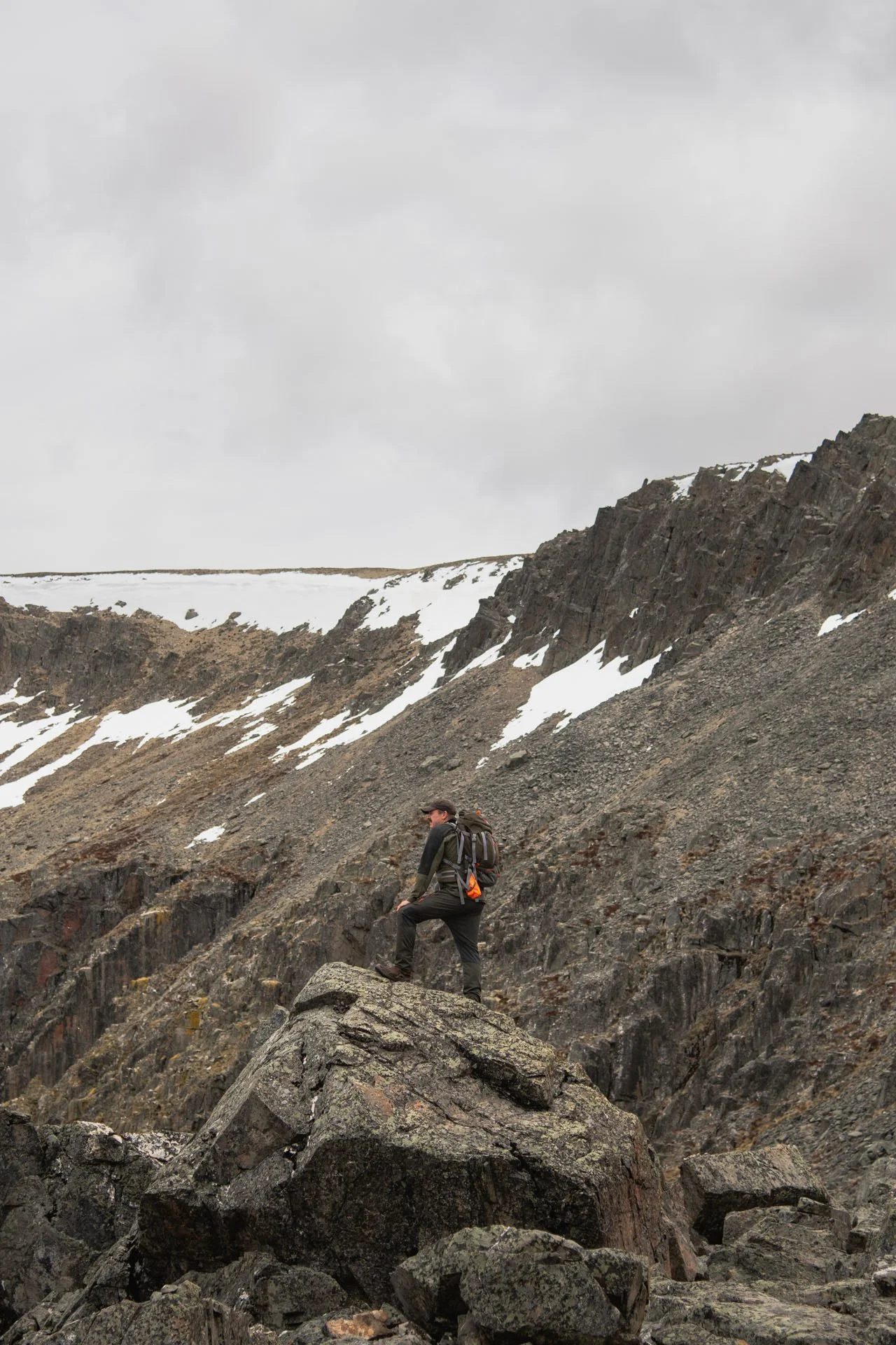 A hiker wearing a backpack and outdoor gear climbing rocky terrain near snow-capped mountains and a cloudy sky.