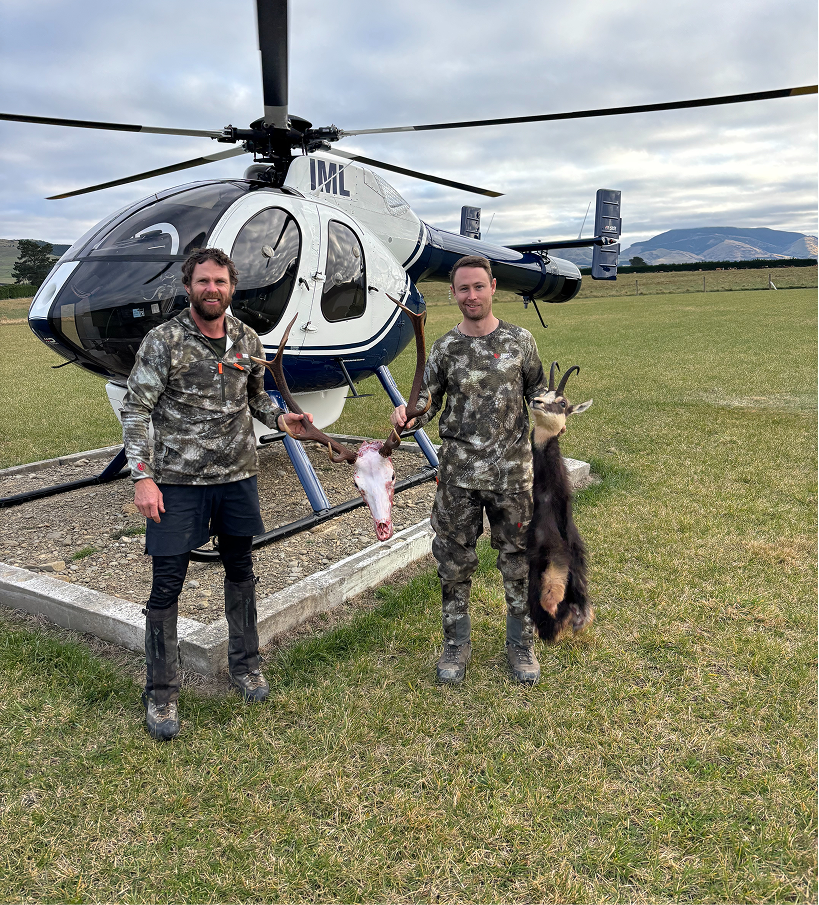 Two men in camouflage clothing standing in front of a helicopter, each holding a hunted animal, with a landscape and mountains in the background.