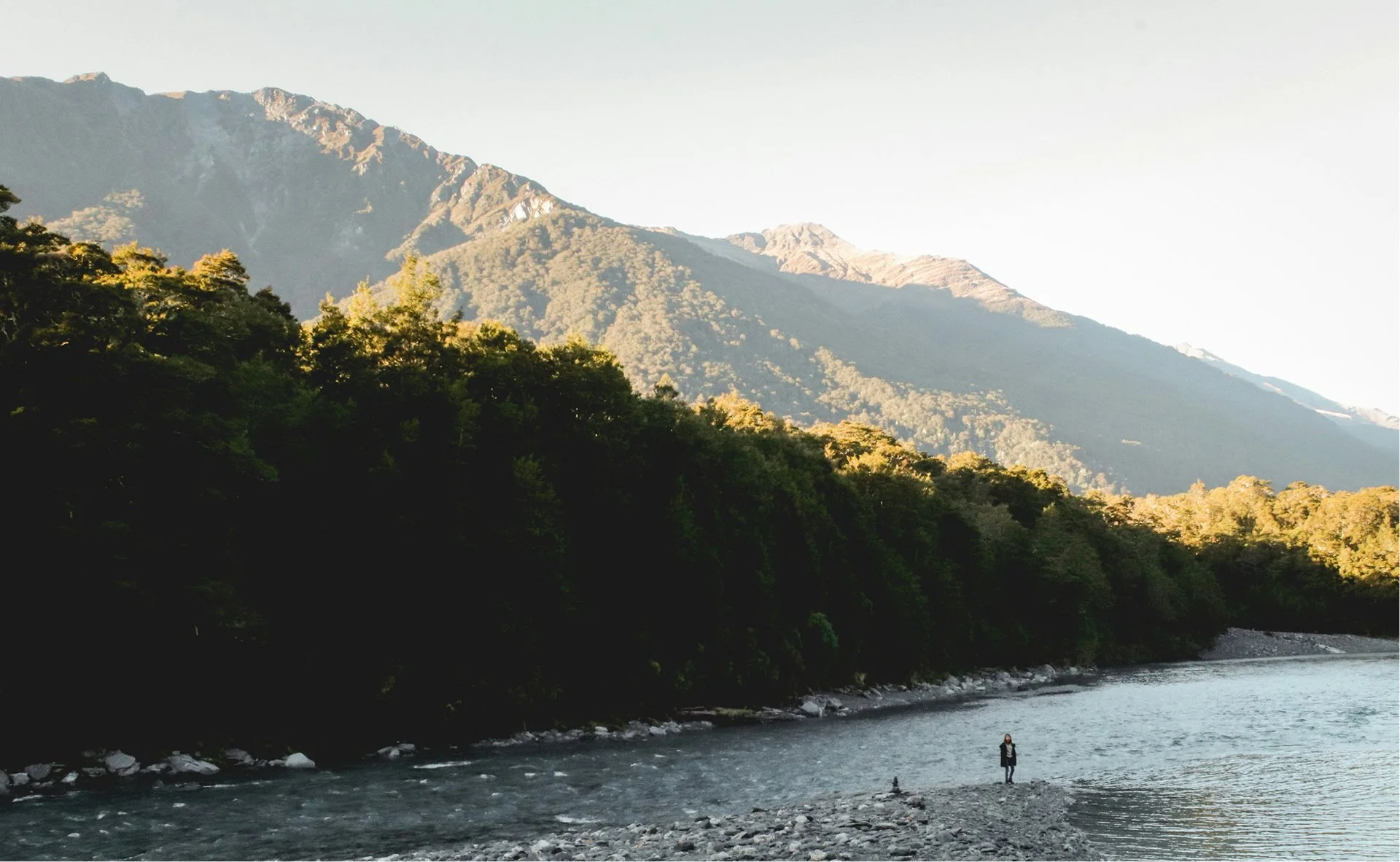 A person standing on a rocky riverbank with a mountain range in the background and trees along the river.
