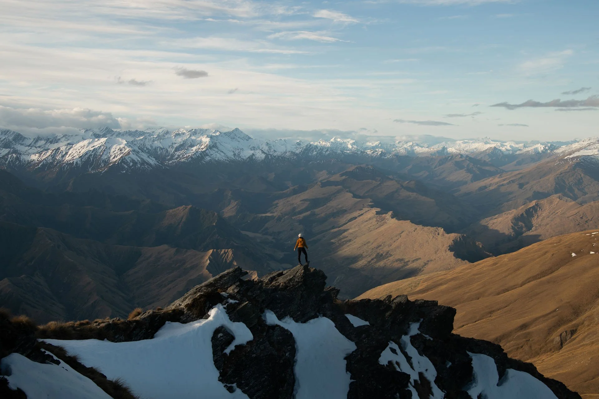 A person wearing a yellow jacket and helmet standing on a rocky mountain ledge overlooking a vast mountain valley with snow-capped peaks and a cloudy sky.