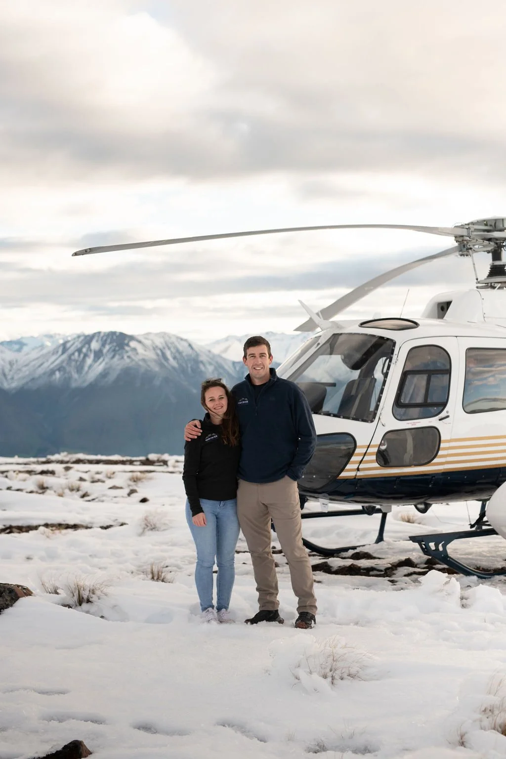 A smiling man and woman standing close together in snowy landscape in front of a helicopter, with snow-capped mountains in the background.