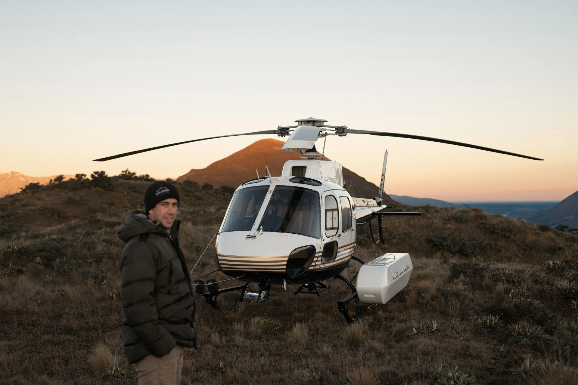 A man in a black jacket and beanie standing next to a white helicopter on a grassy hillside with mountains in the background during sunset.