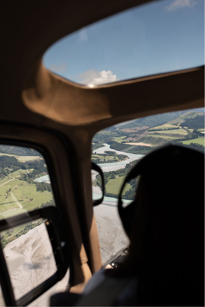 A view from inside a helicopter showing a river winding through green fields and hills, with a sky and clouds visible through the window.