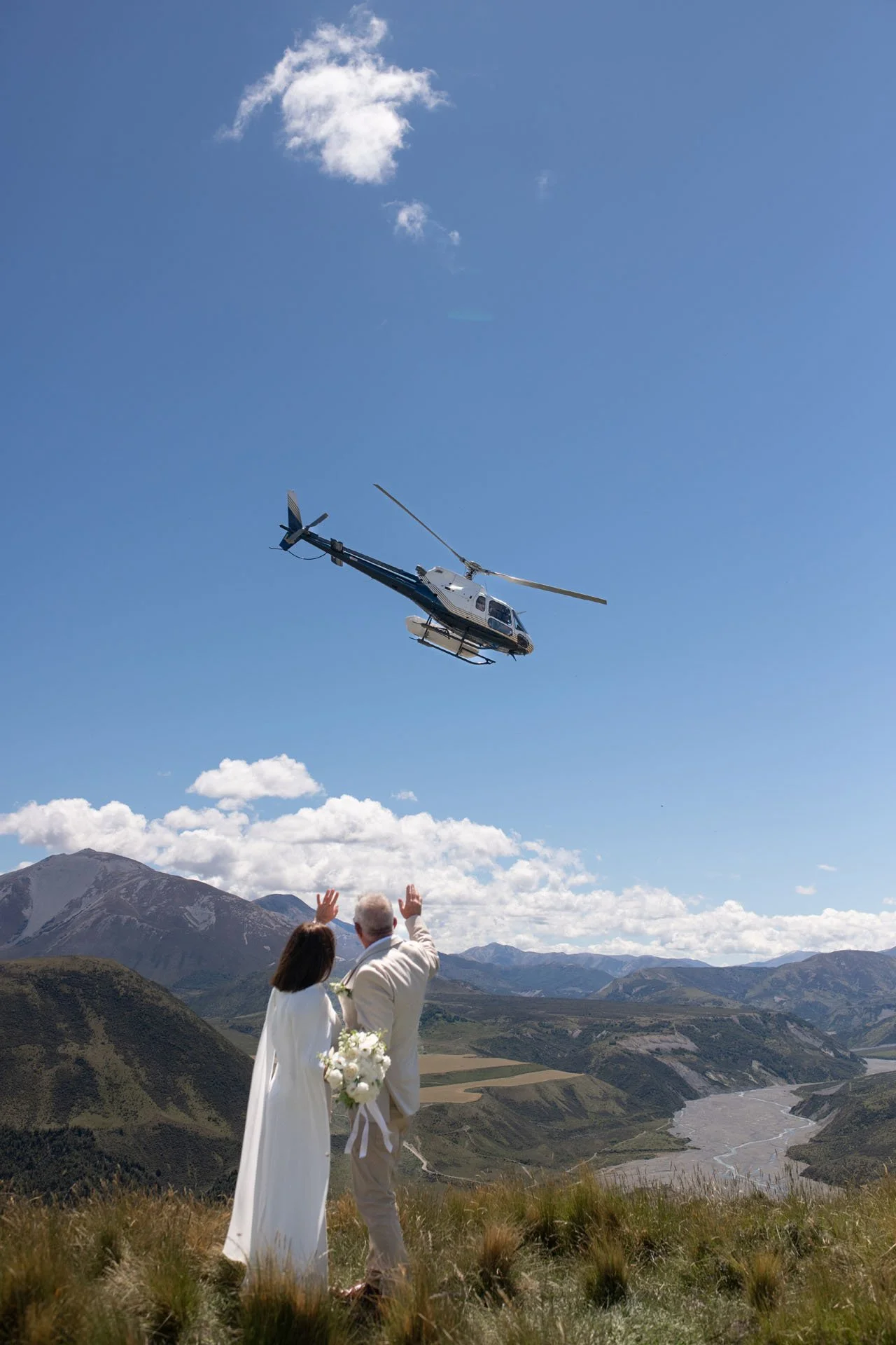 A couple is celebrating a wedding outdoors in a mountainous landscape with a helicopter flying overhead. The bride is wearing a white wedding dress and holding a bouquet, and the groom is dressed in a light-colored suit. They are looking up at the helicopter, which is flying above them against a bright blue sky, with rocky mountains and a river in the background.