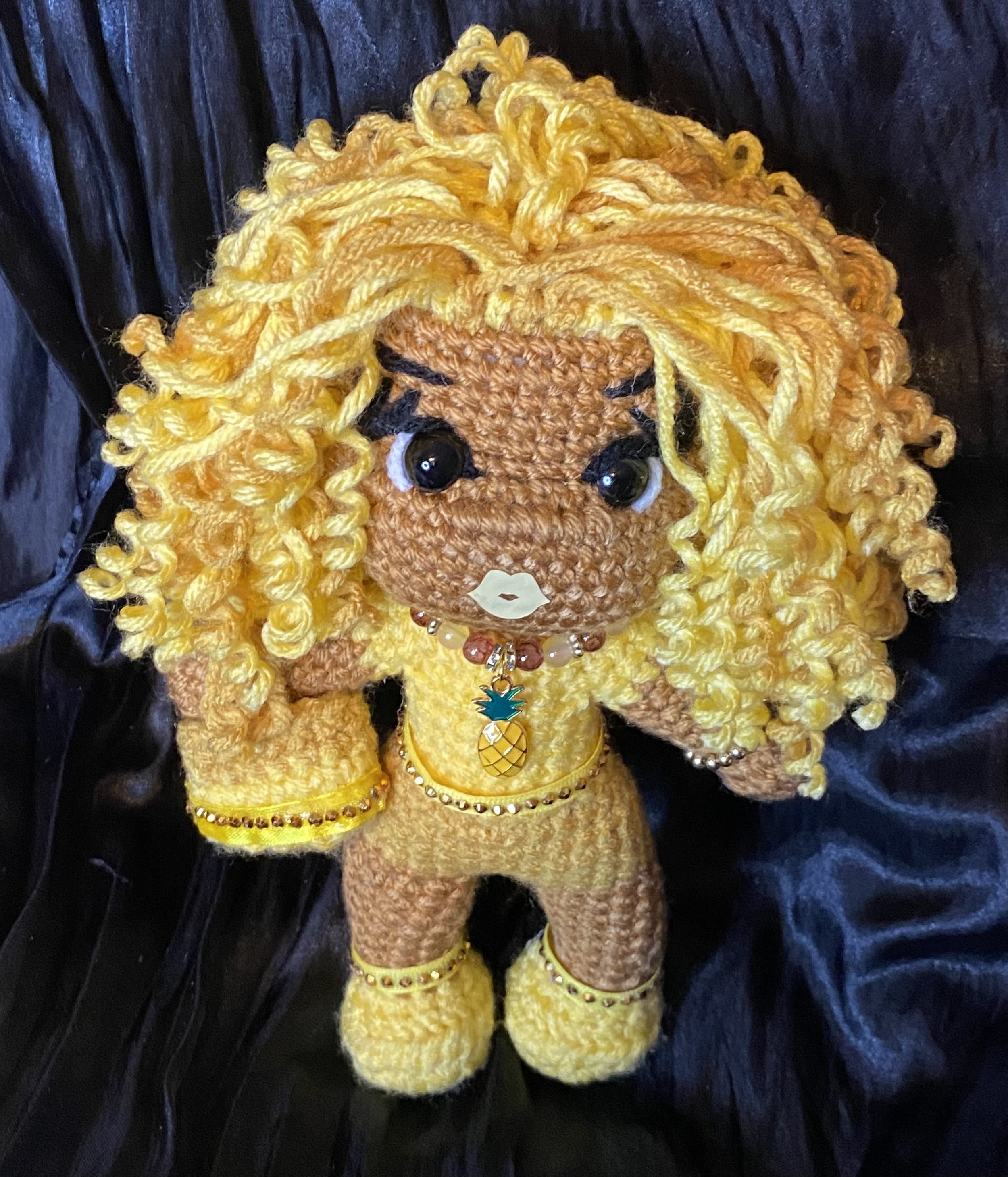 Handmade crocheted doll in a yellow dress with pineapple-shaped pendant, with curly yellow hair, black eyes, and jewelry, lying on a black surface.