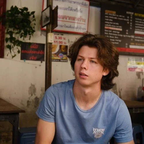 Young man with wavy brown hair wearing a light blue t-shirt sitting in a restaurant or café