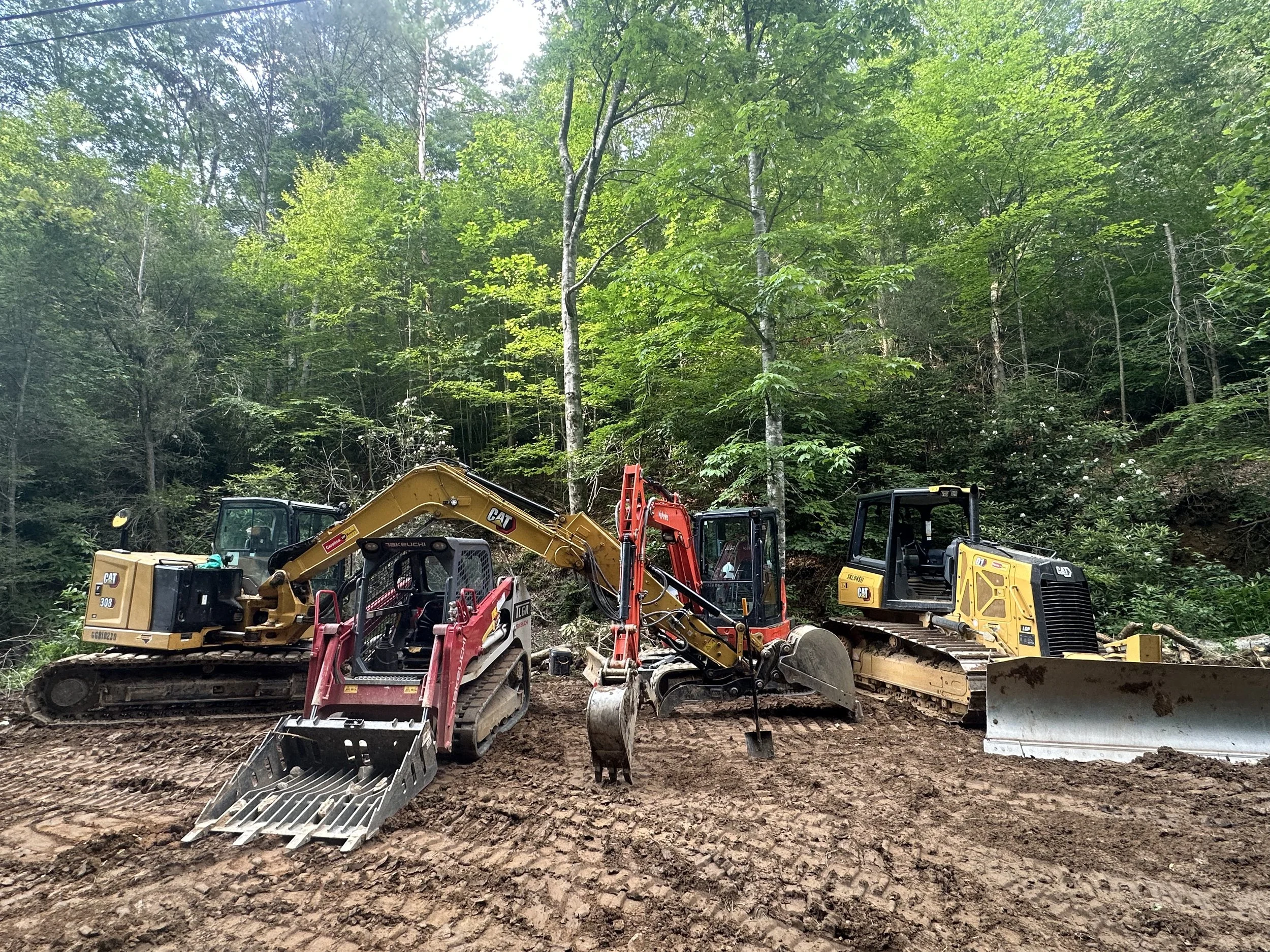 Construction equipment, including excavators, bulldozers, and a roller, positioned on uneven, disturbed dirt ground in a forested area with green trees in the background.
