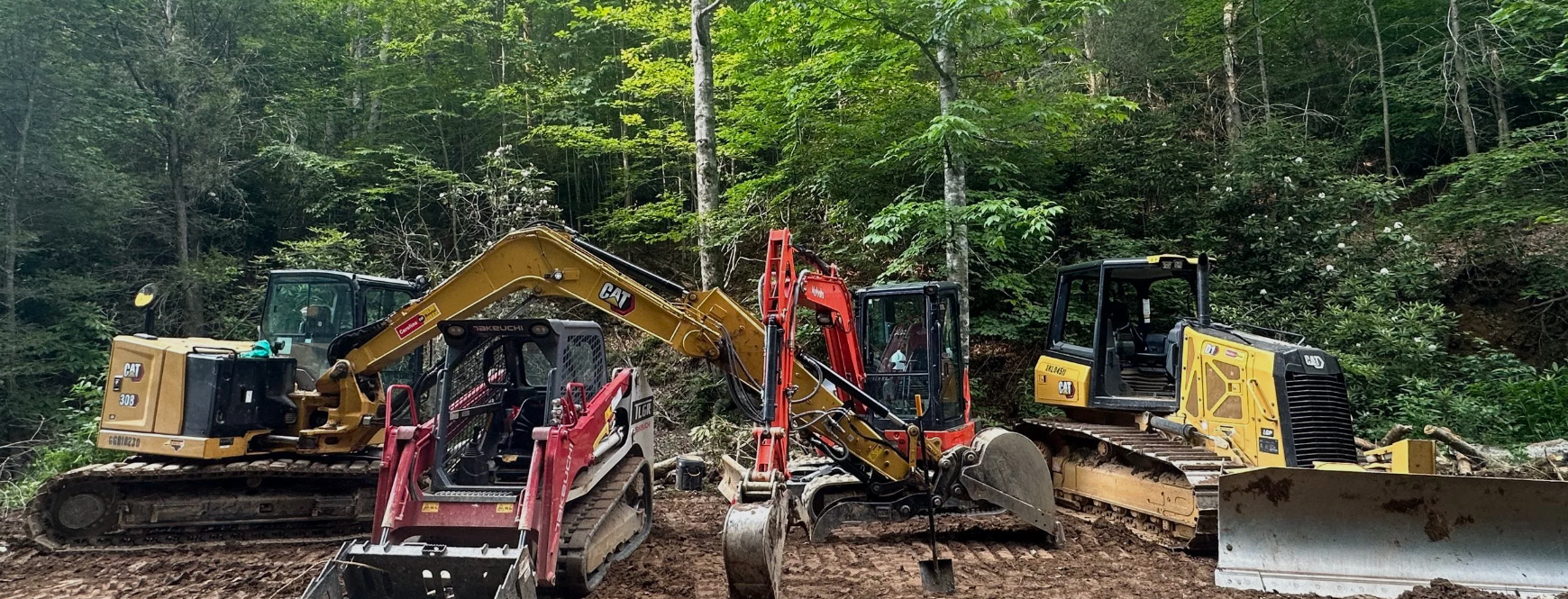 Three pieces of construction equipment, including a yellow CAT excavator, a black and red Takeuchi mini excavator, and a yellow CAT bulldozer, are positioned in a forested area with green trees and foliage.