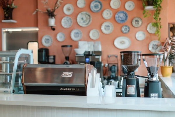Coffee shop countertop with a G2 espresso machine, coffee grinder, and cups, with decorative plates on the wall behind.