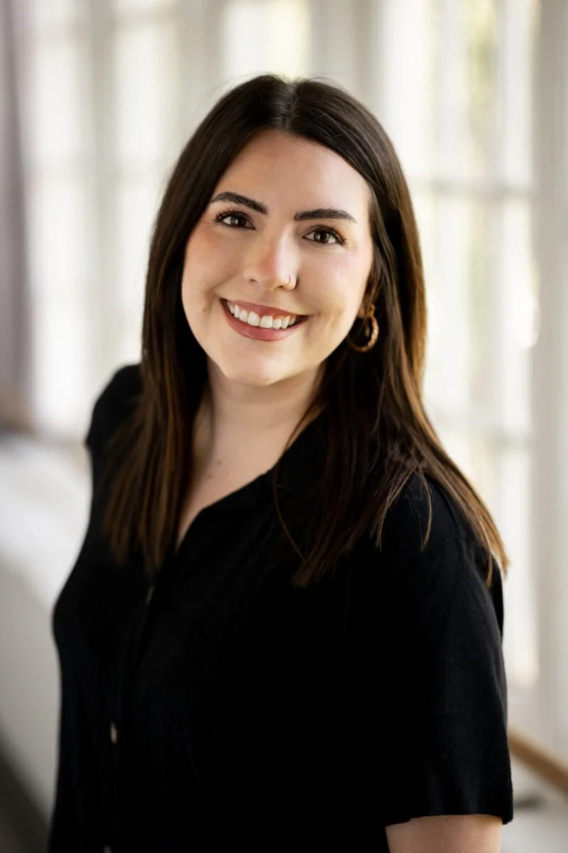A woman with long dark hair smiling, wearing a black top and gold hoop earrings, standing in a bright room with large windows and white curtains.