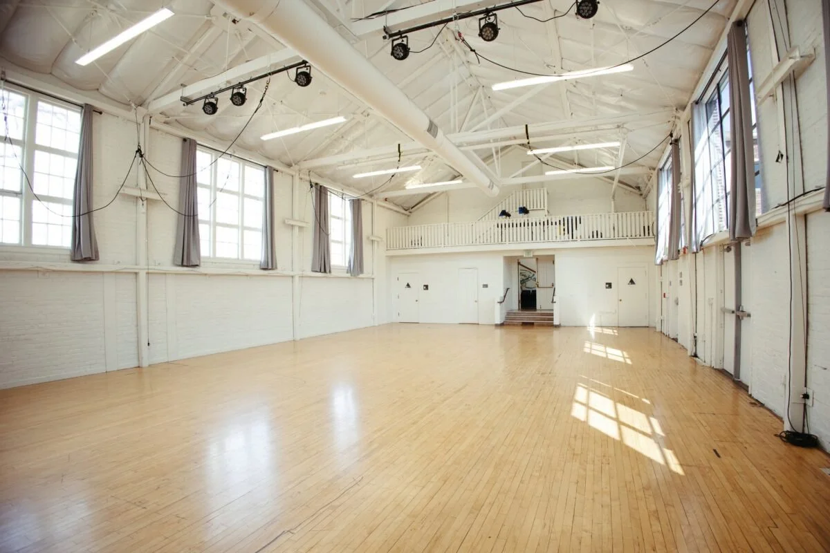 An empty dance or event hall with hardwood floors, white brick walls, large windows with curtains, and a high ceiling with exposed beams and lighting.